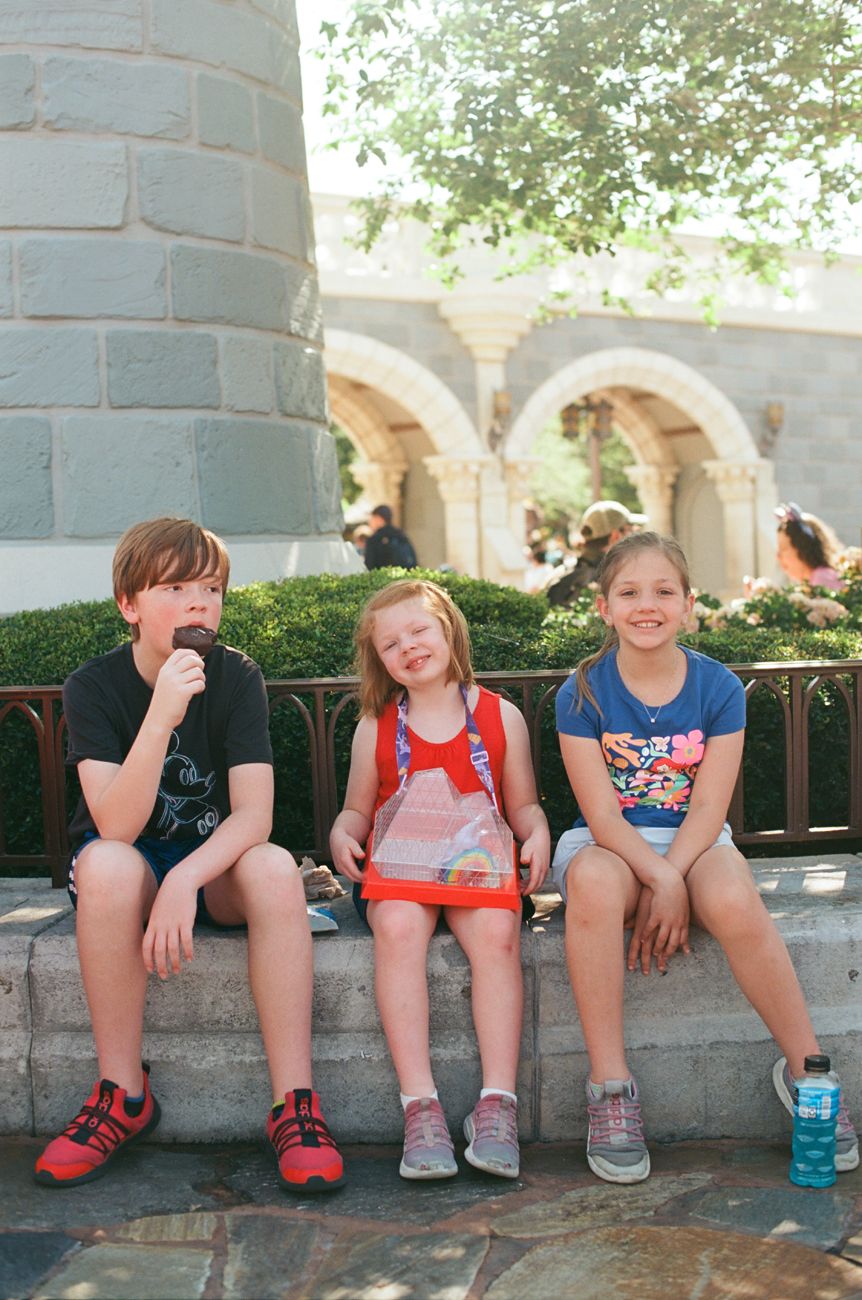 Three young visitors relaxing on a stone wall near a castle-like structure at Disney World.