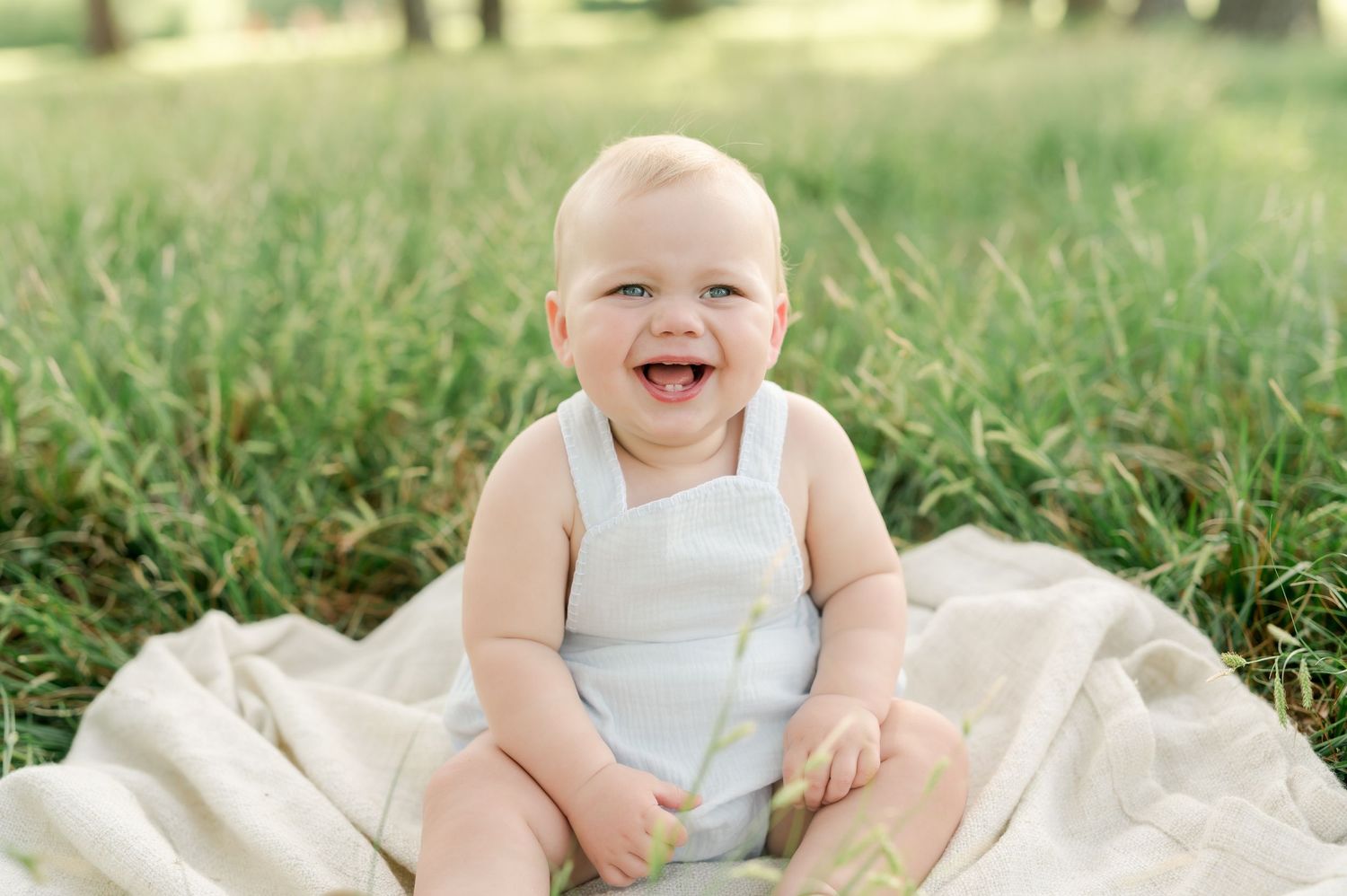 Baby in white clothing sits on cream blanket outdoors in natural sunlit setting.