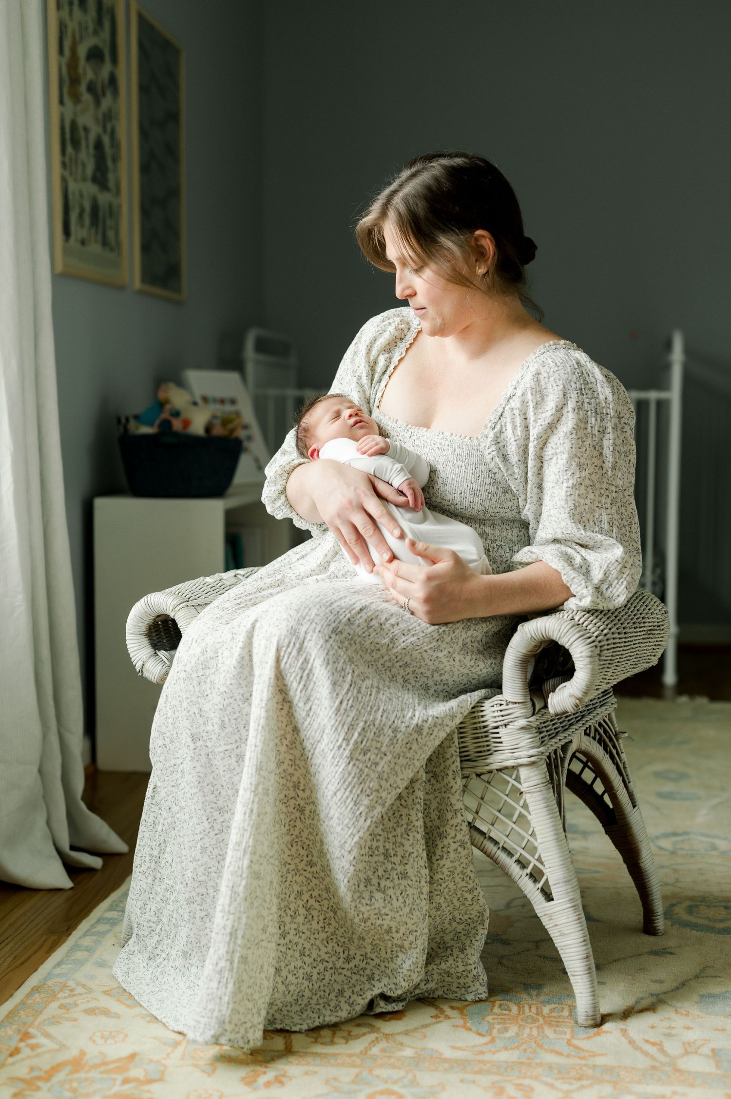 Person in flowing white dress sits in rocking chair holding infant in bright indoor setting.