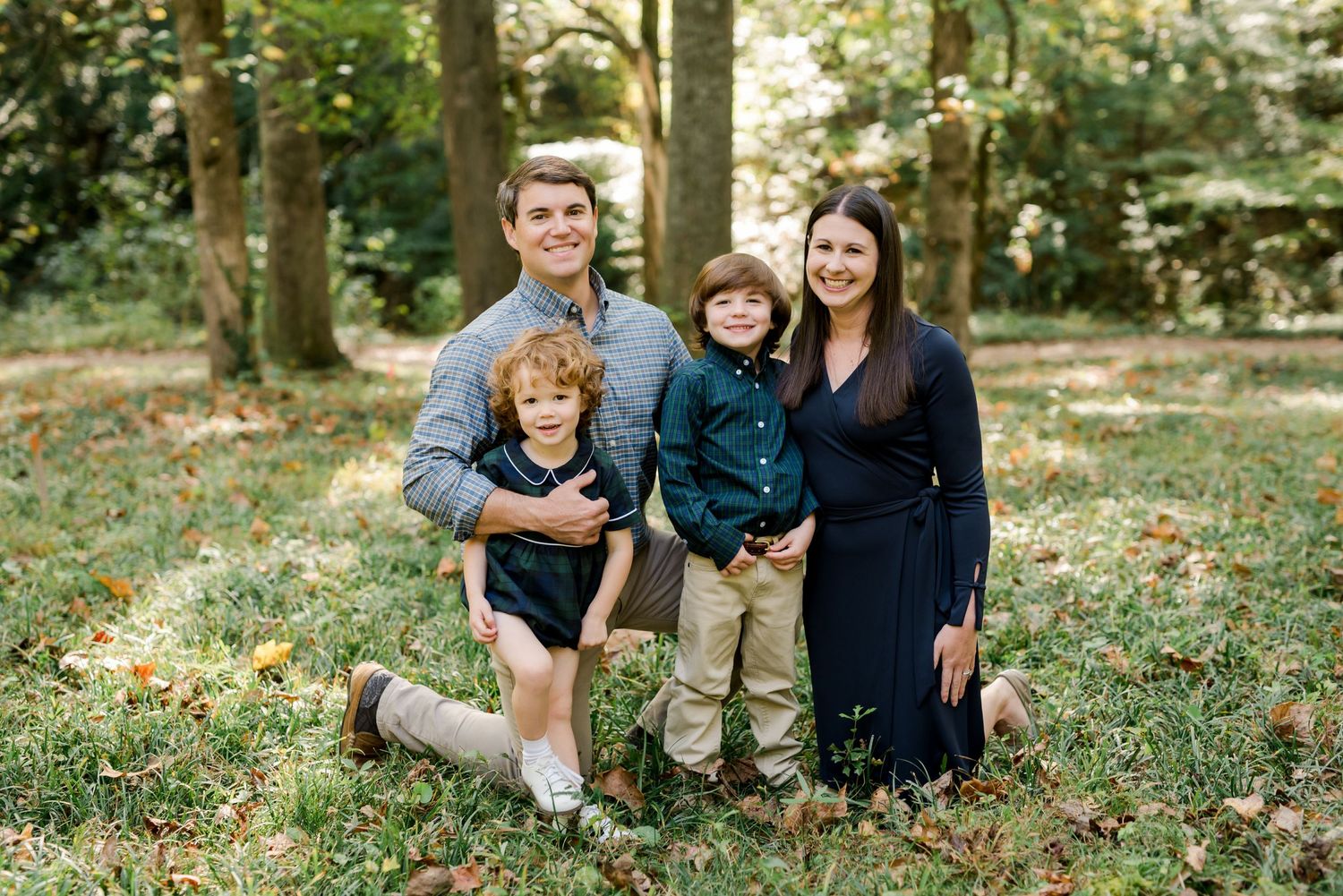 Family of four poses together on fallen leaves in a wooded setting wearing coordinated navy and neutral outfits.