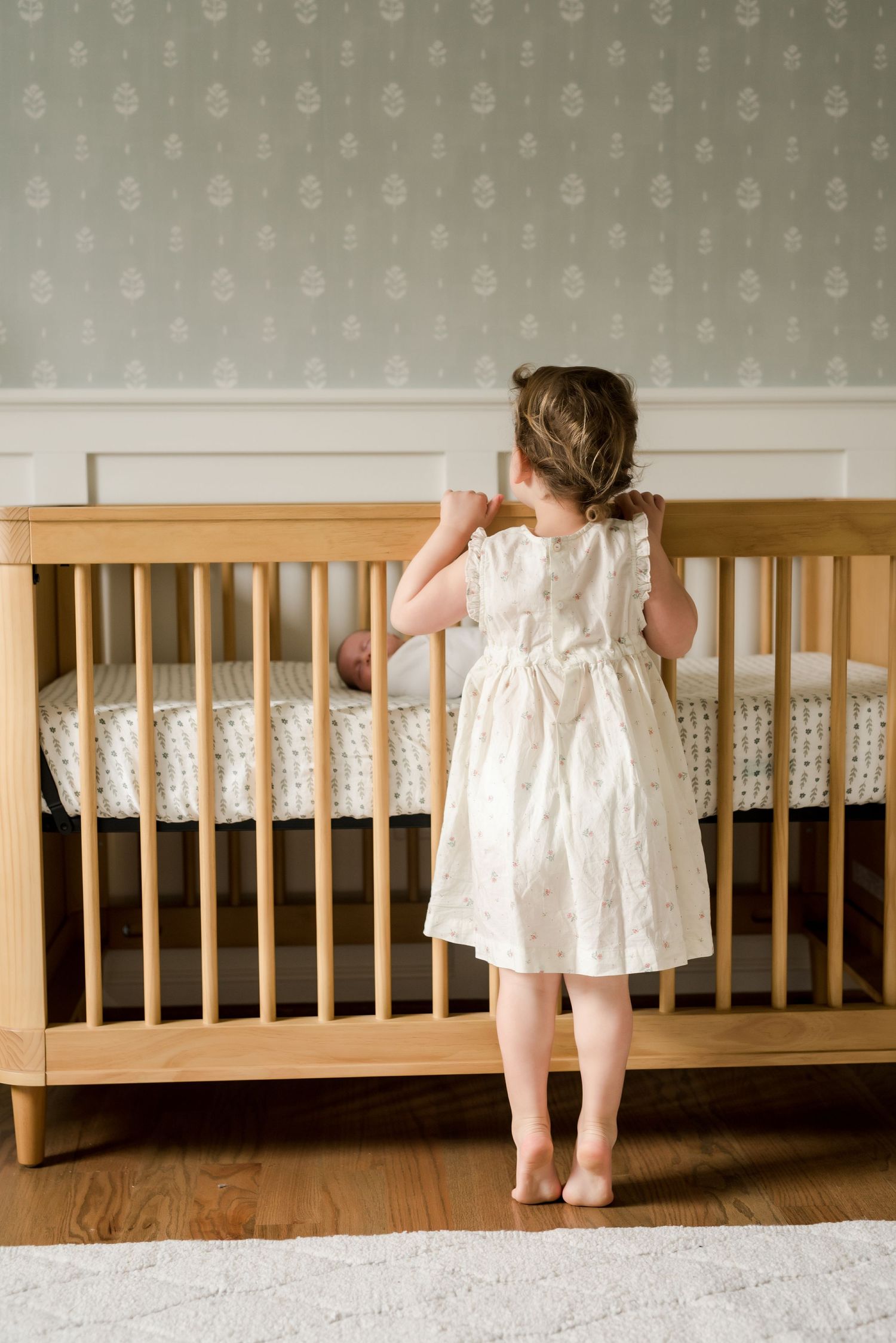 A small child in a white dress stands beside a wooden crib looking over the railing in a nursery room.
