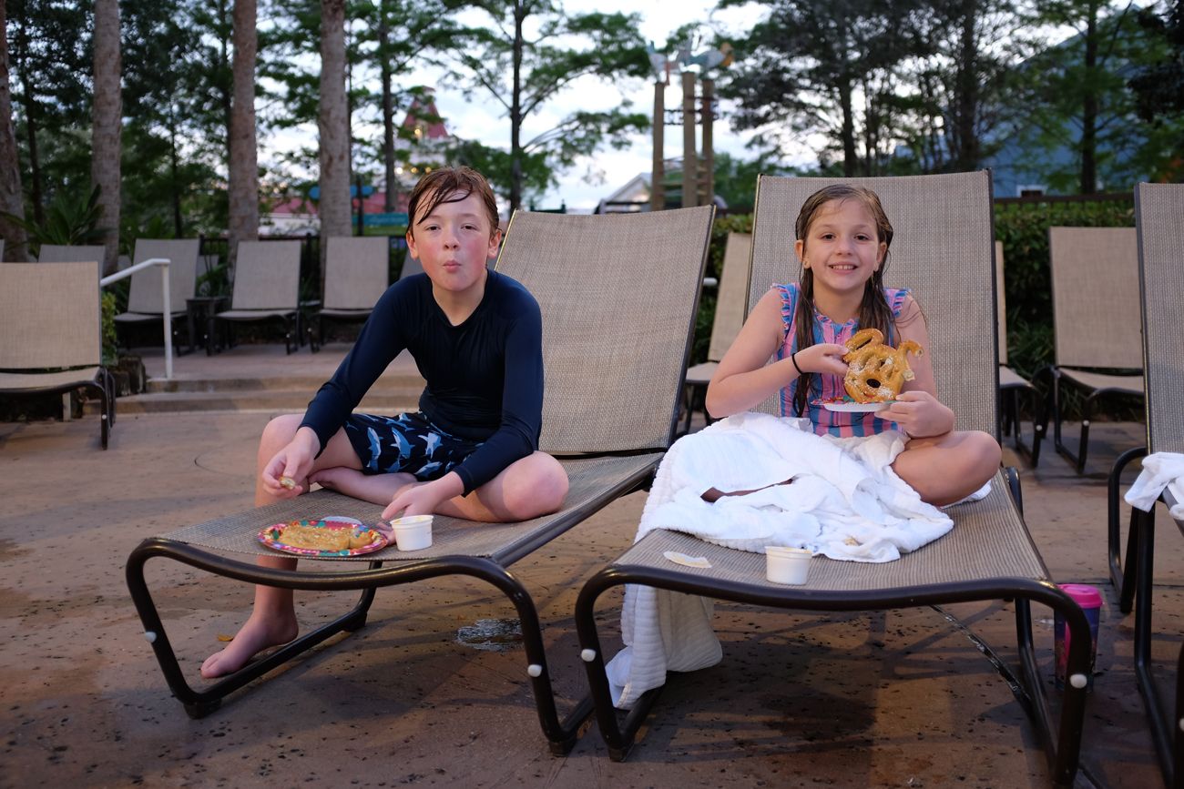 Two people relaxing on lounge chairs at poolside area surrounded by pine trees.