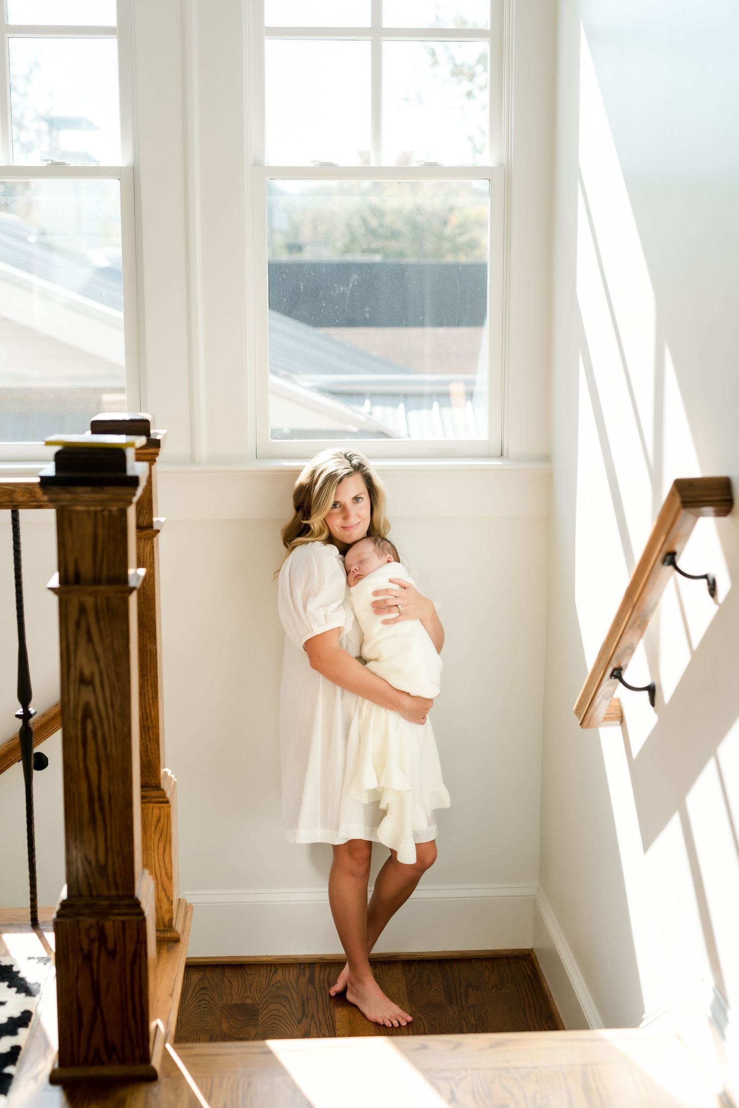 Woman in white dress holds baby in bright stairwell with natural light streaming through windows.