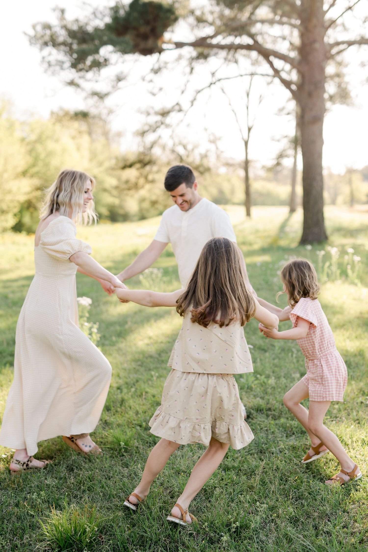 A family holds hands while dancing in a circle in a sunlit meadow wearing neutral tones.