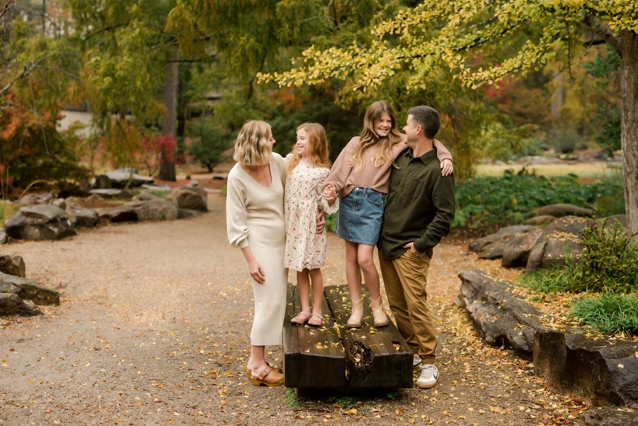 Family of four laughing together on a garden path in autumn, with a vintage suitcase at their feet.