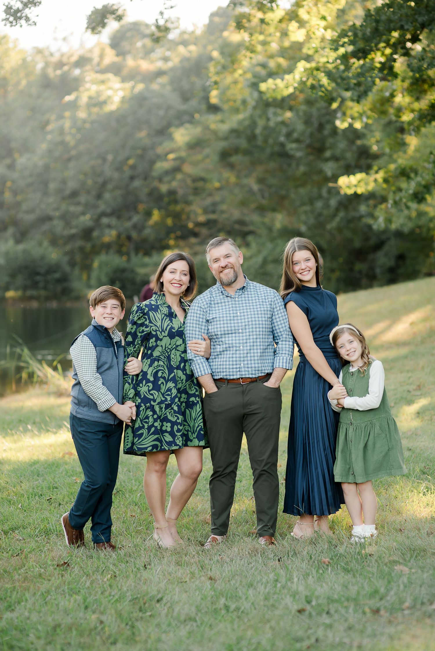 Family of five smiles together on a grassy lawn surrounded by trees in coordinating green and navy outfits.