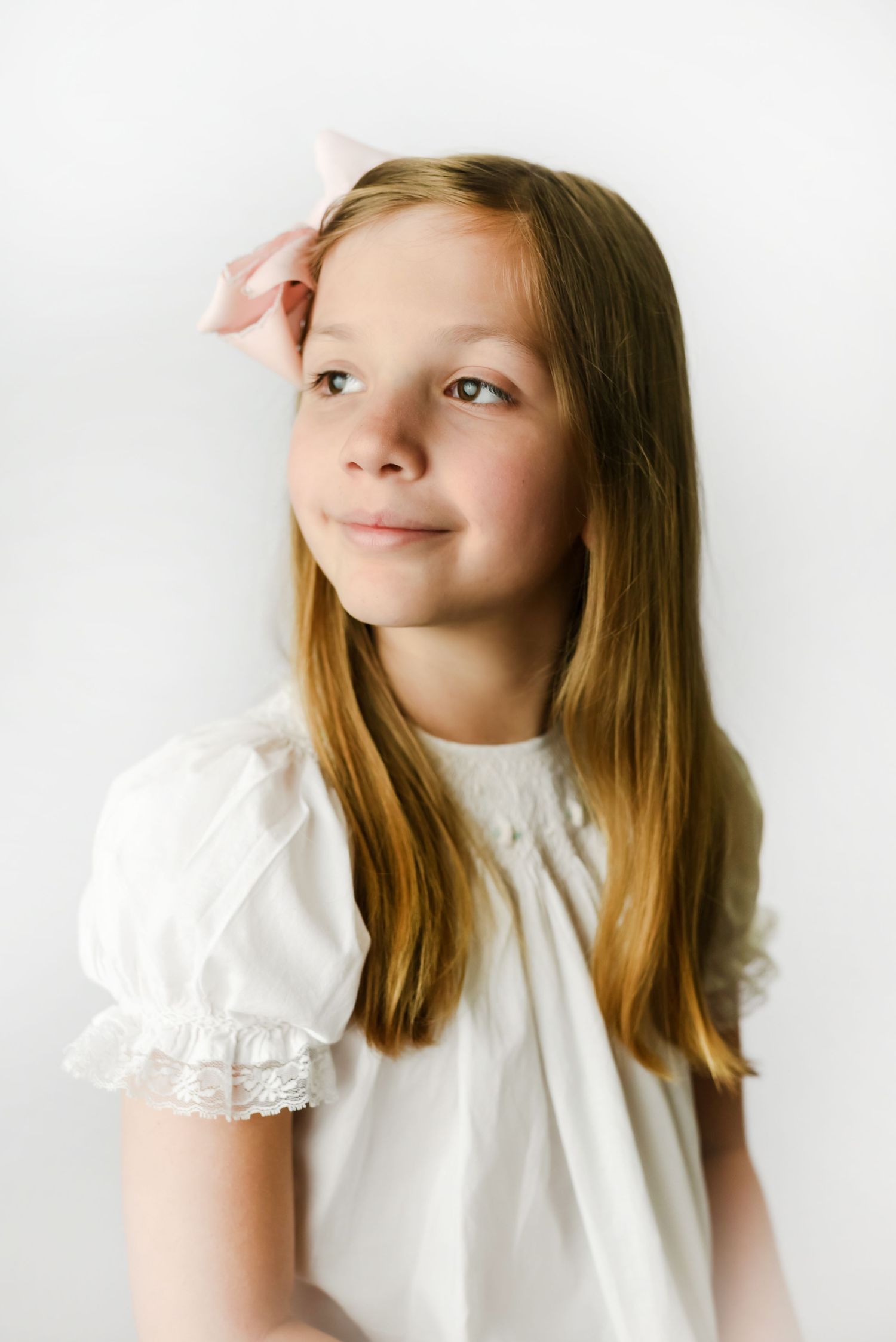 Young person in white lace-trimmed dress poses against white background with gentle smile.