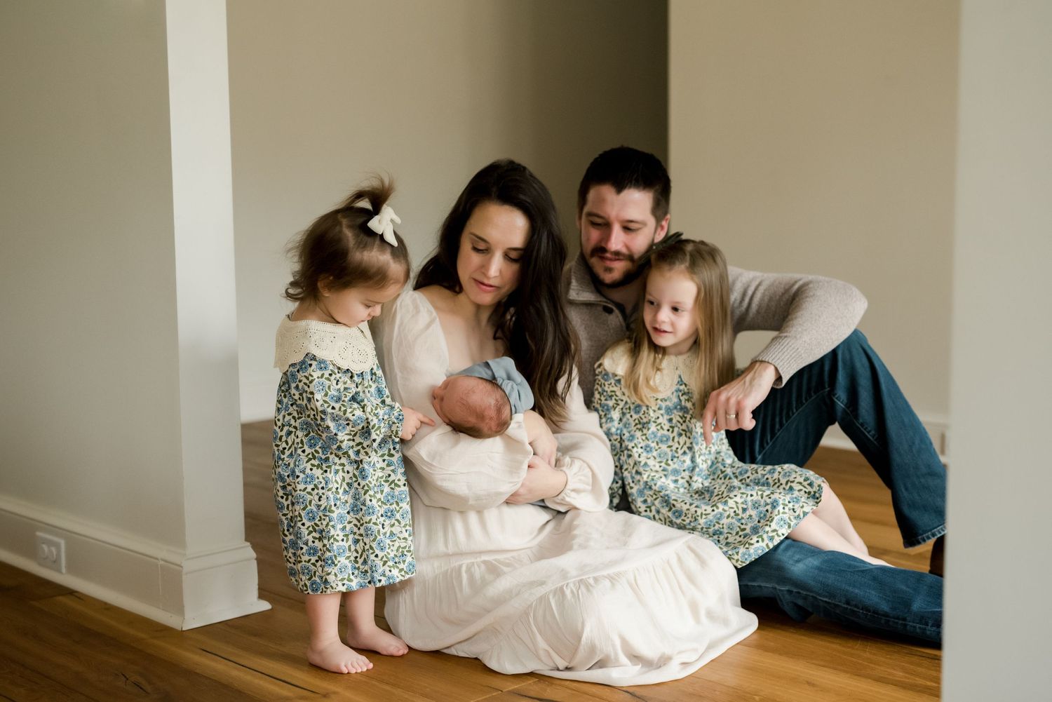 Family gathered on hardwood floor with newborn baby in indoor setting.