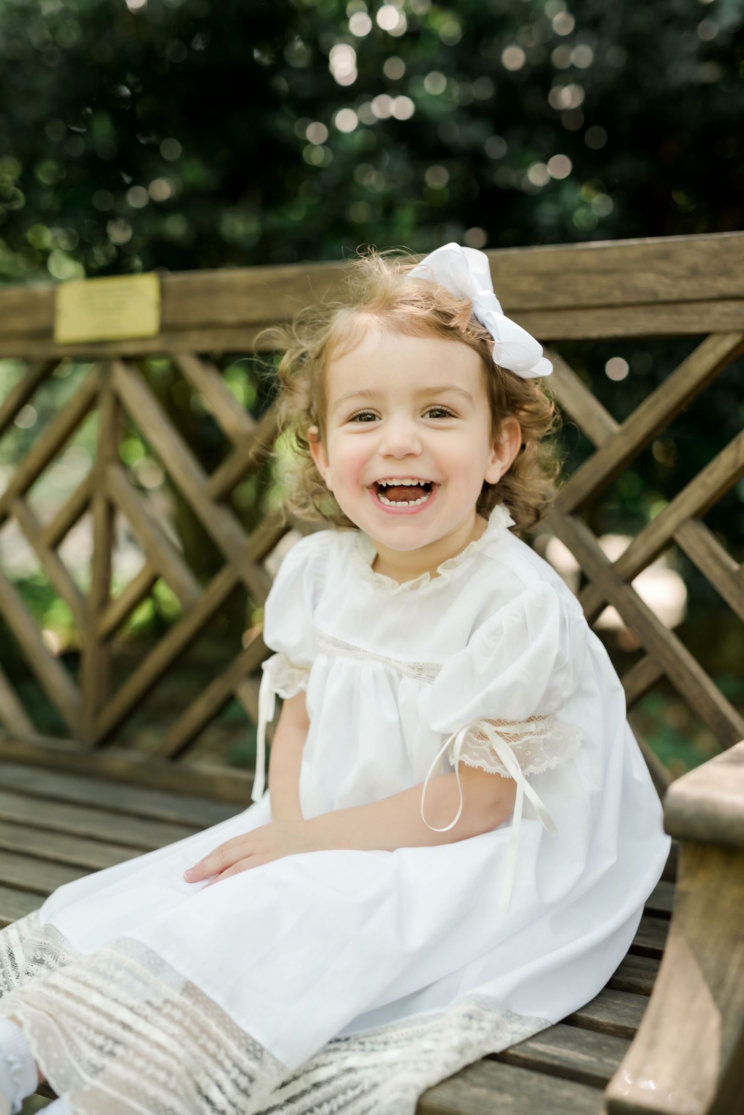 A happy child in a white dress with a bow sits on a wooden bench outdoors.