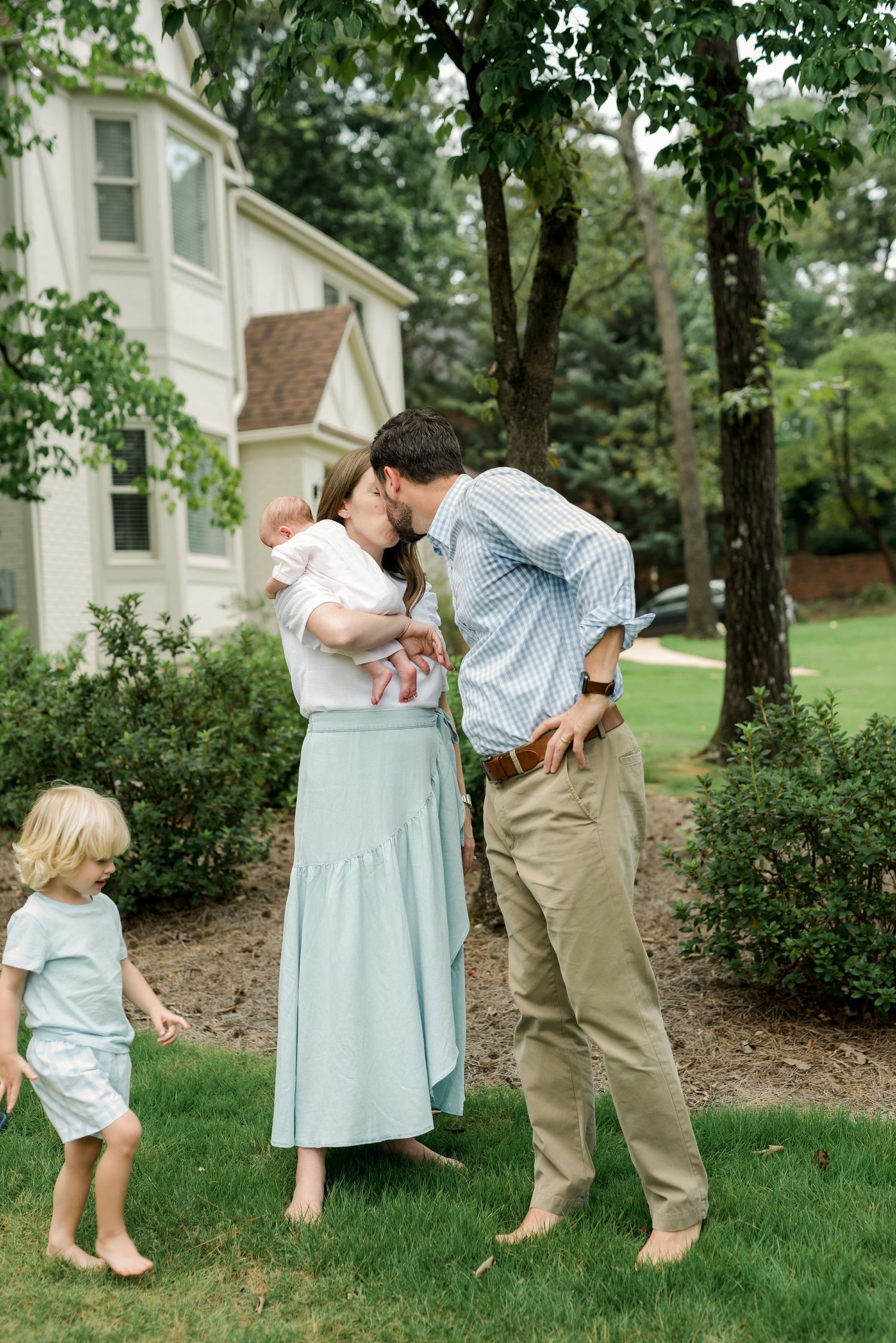 Playful family moment on lawn with white house and trees in background.