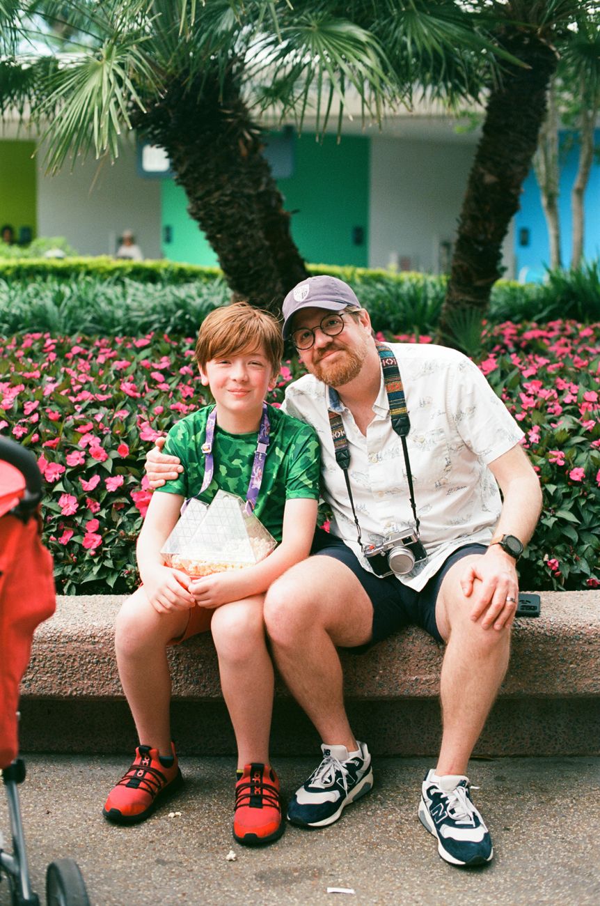 Two visitors sitting on a stone bench surrounded by tropical plants and pink flowers at a theme park.
