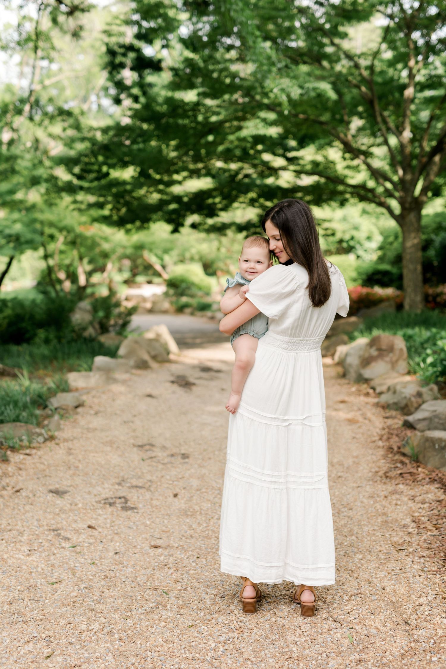 Woman in white dress holding baby while standing on garden path surrounded by greenery.