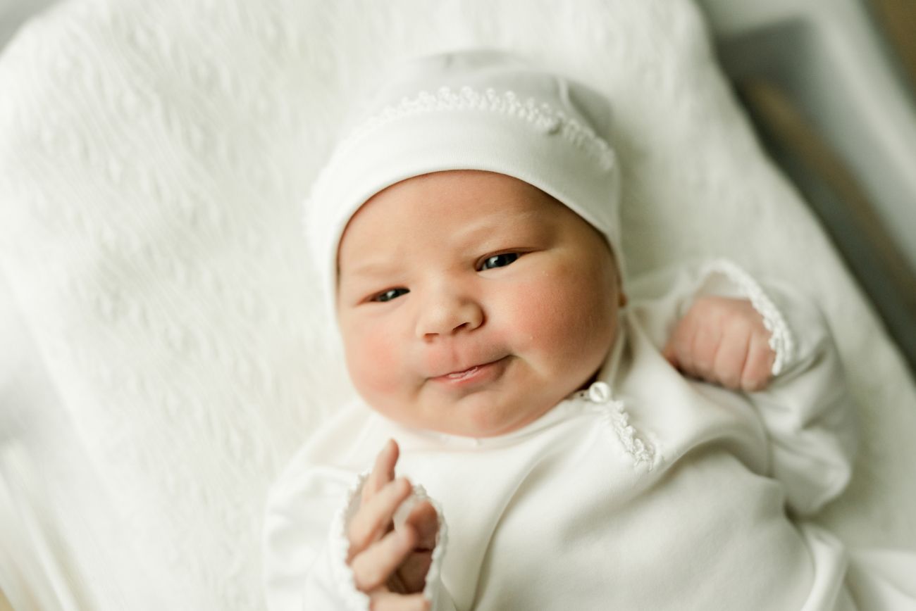 Newborn baby wearing white bonnet and wrap lies peacefully against white background.