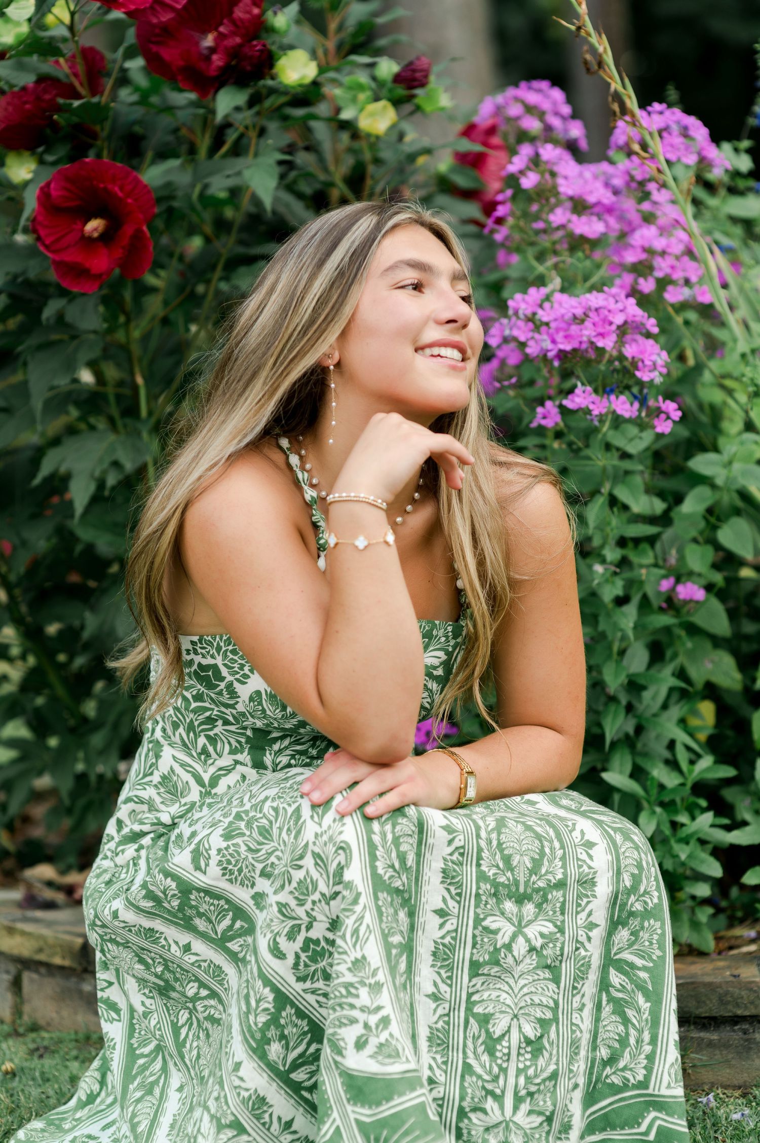 Woman in green floral dress poses among vibrant red and pink flowers in a garden setting.