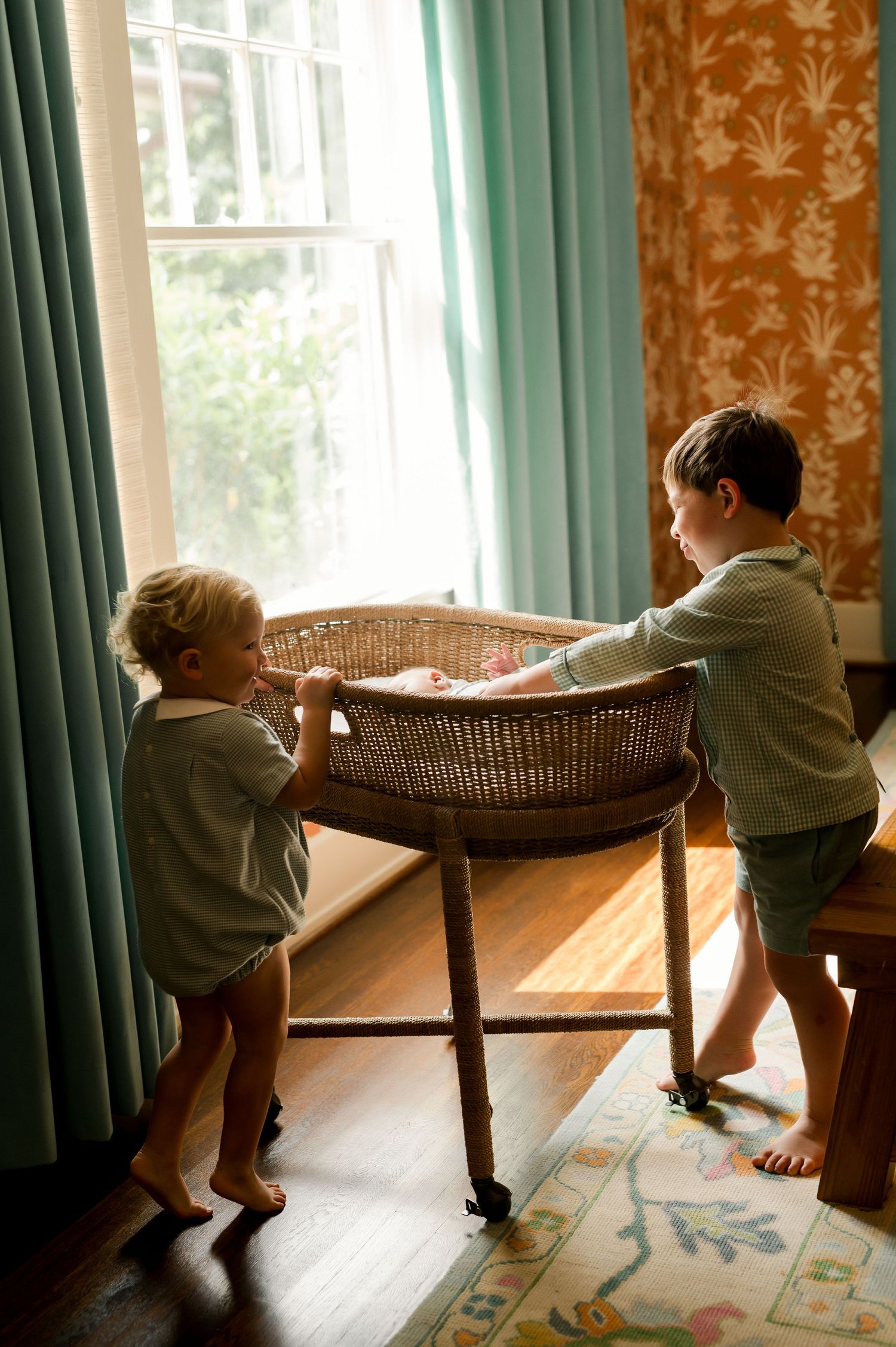 Two young boys stand beside a wicker bassinet with a newborn baby inside, near a sunlit window with teal curtains.