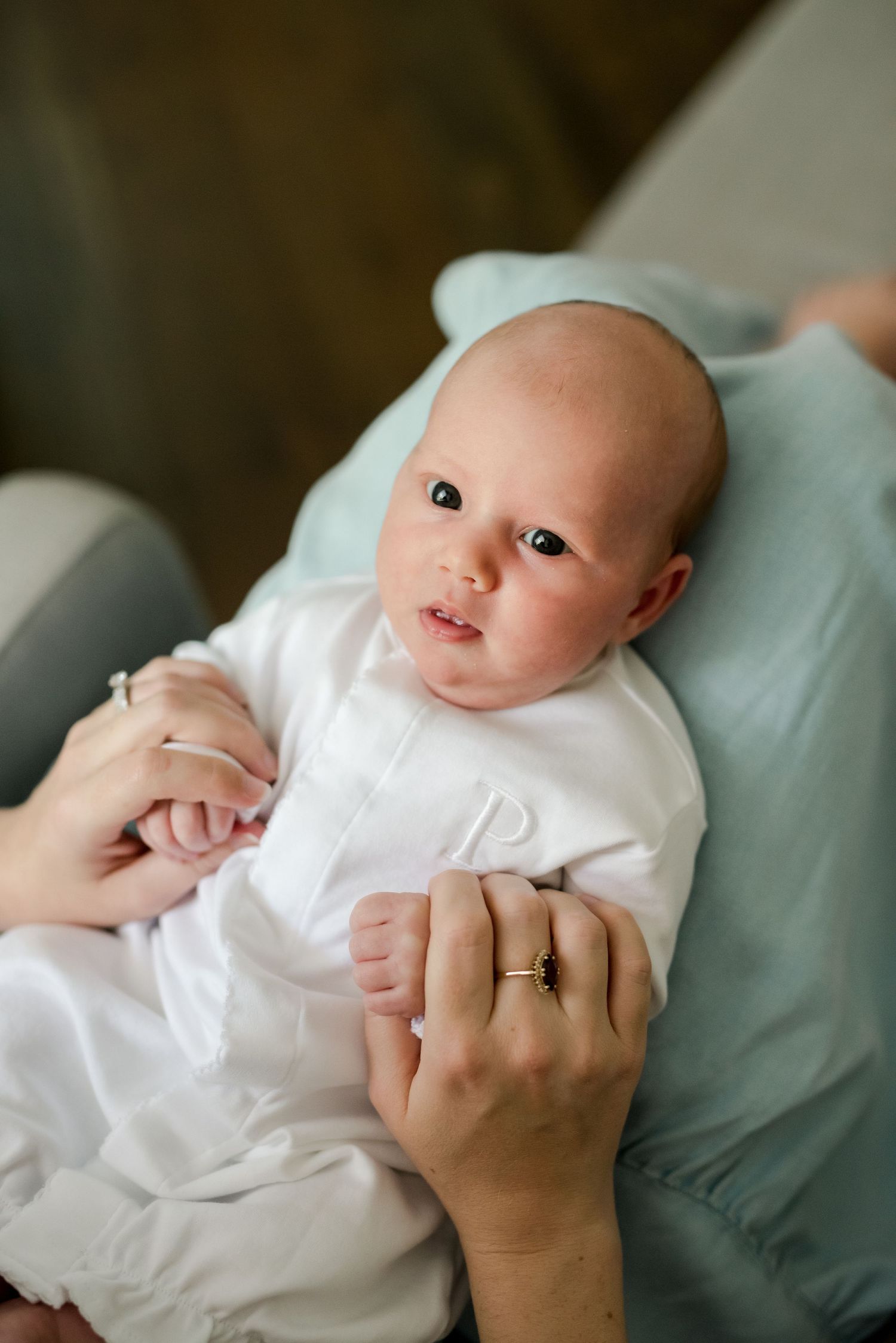 Baby in white christening gown lying on blue cushion with hands being held.