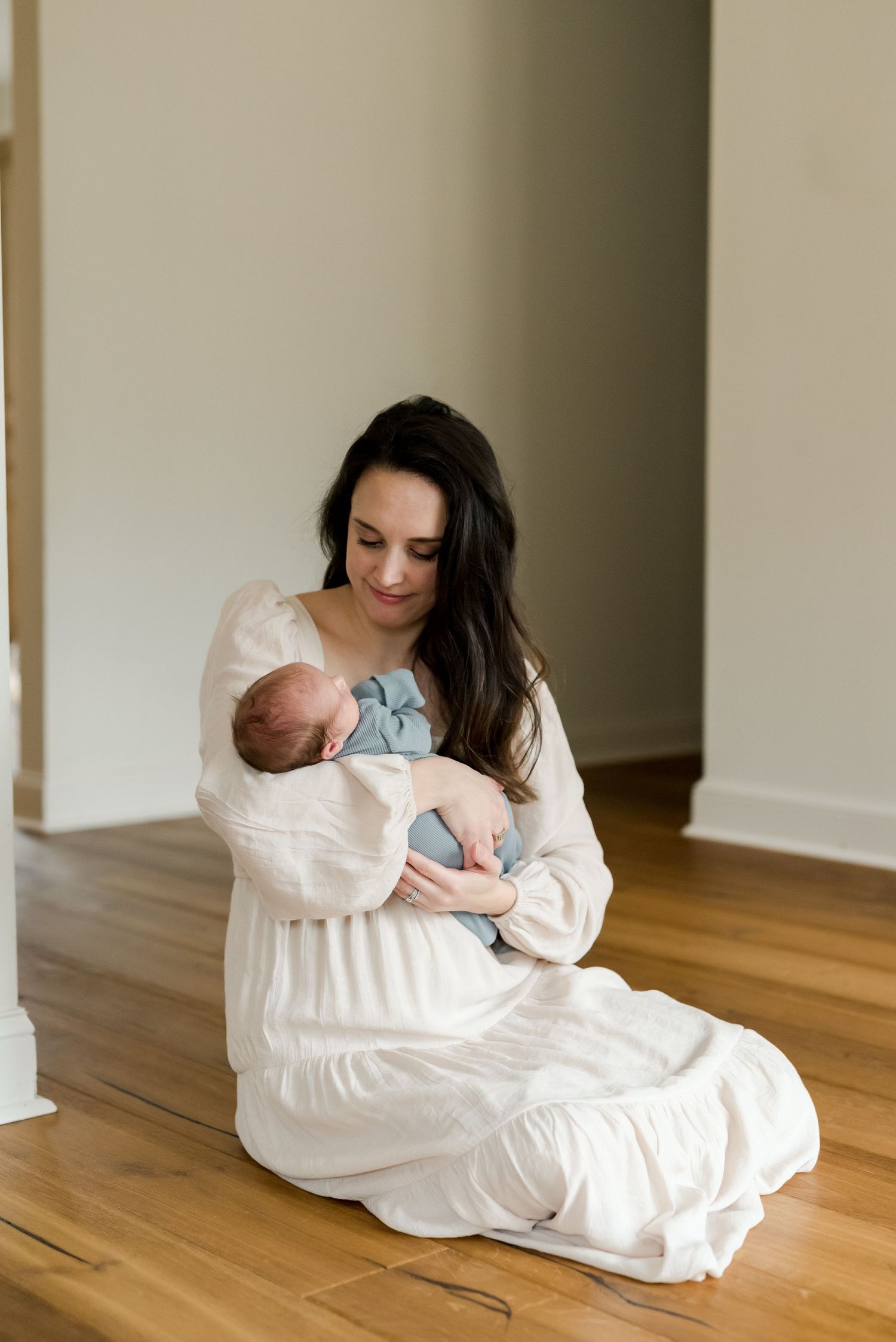 Mother in white dress sitting on hardwood floor holding newborn baby indoors.