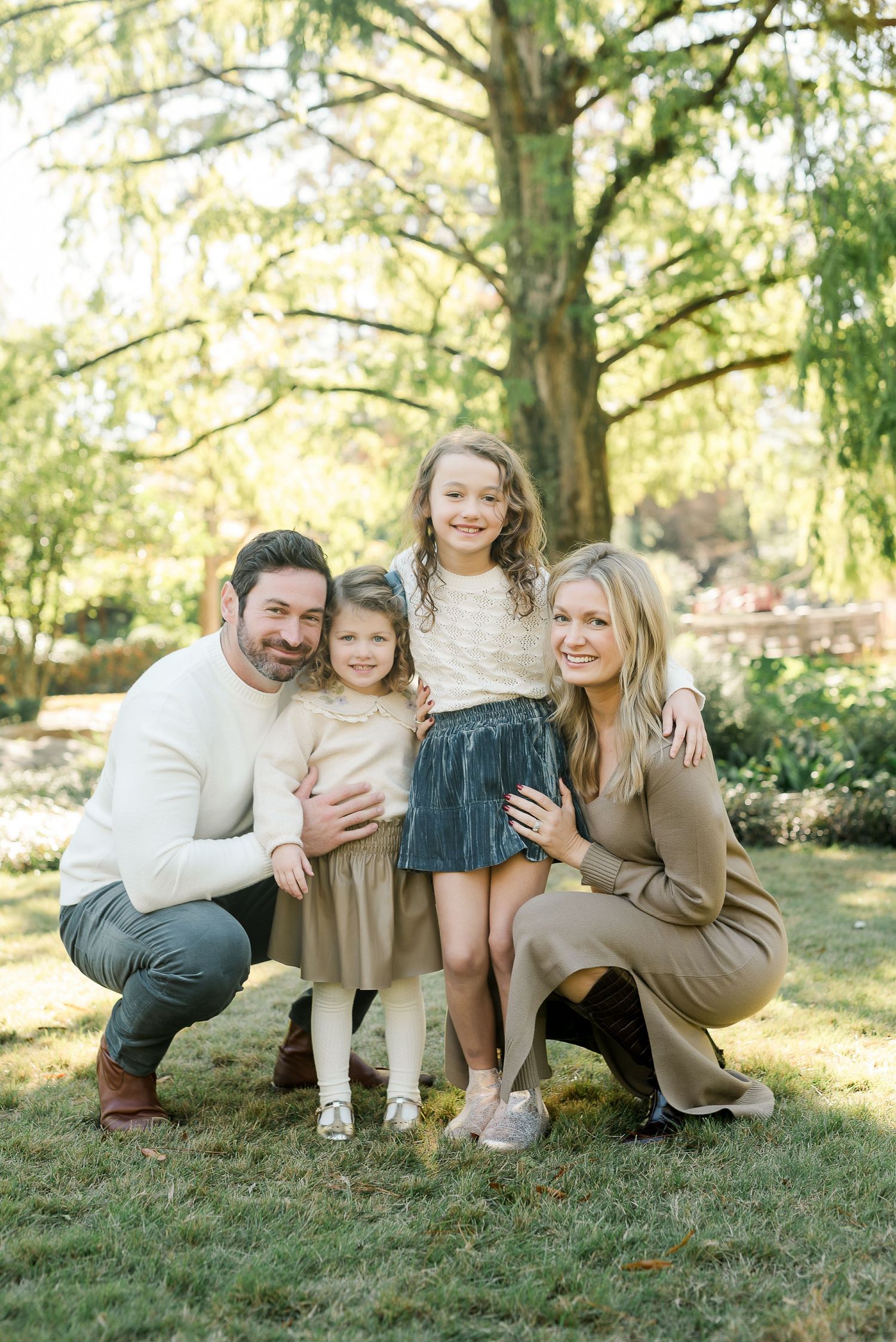 Family of four poses outdoors in neutral tones, parents crouching beside two young daughters under a large tree.