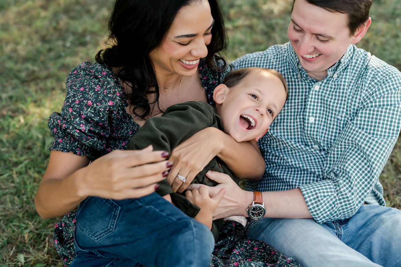 A joyful family of three laughing together outdoors, parents playfully holding their giggling toddler son.
