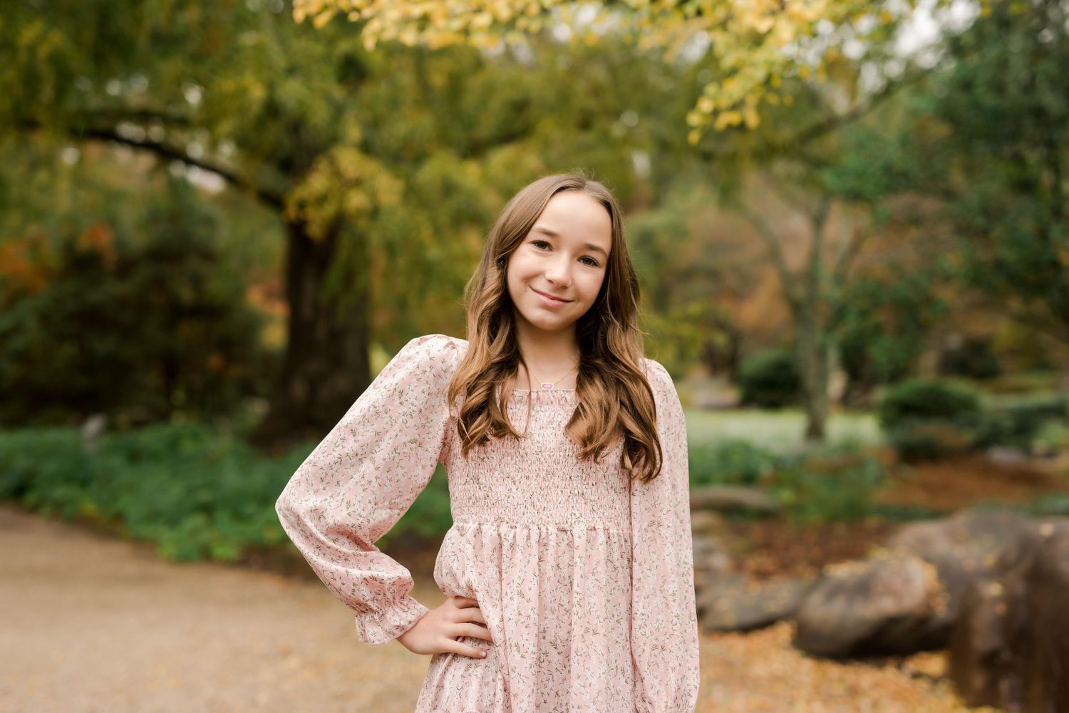 Young person in a floral dress poses in a garden with fall foliage in the background.