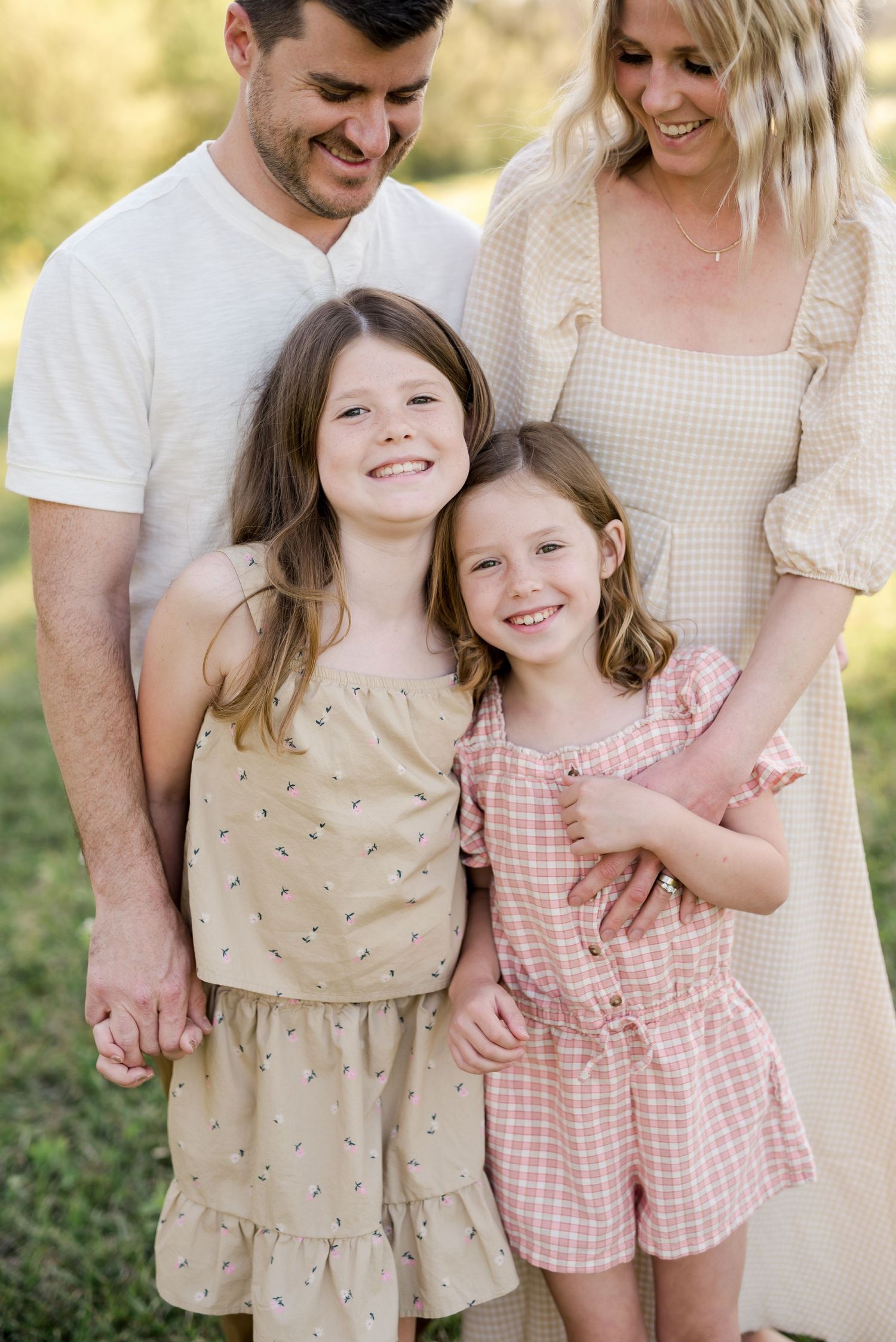 A family of four wearing coordinating neutral outfits poses together outdoors in golden sunlight.