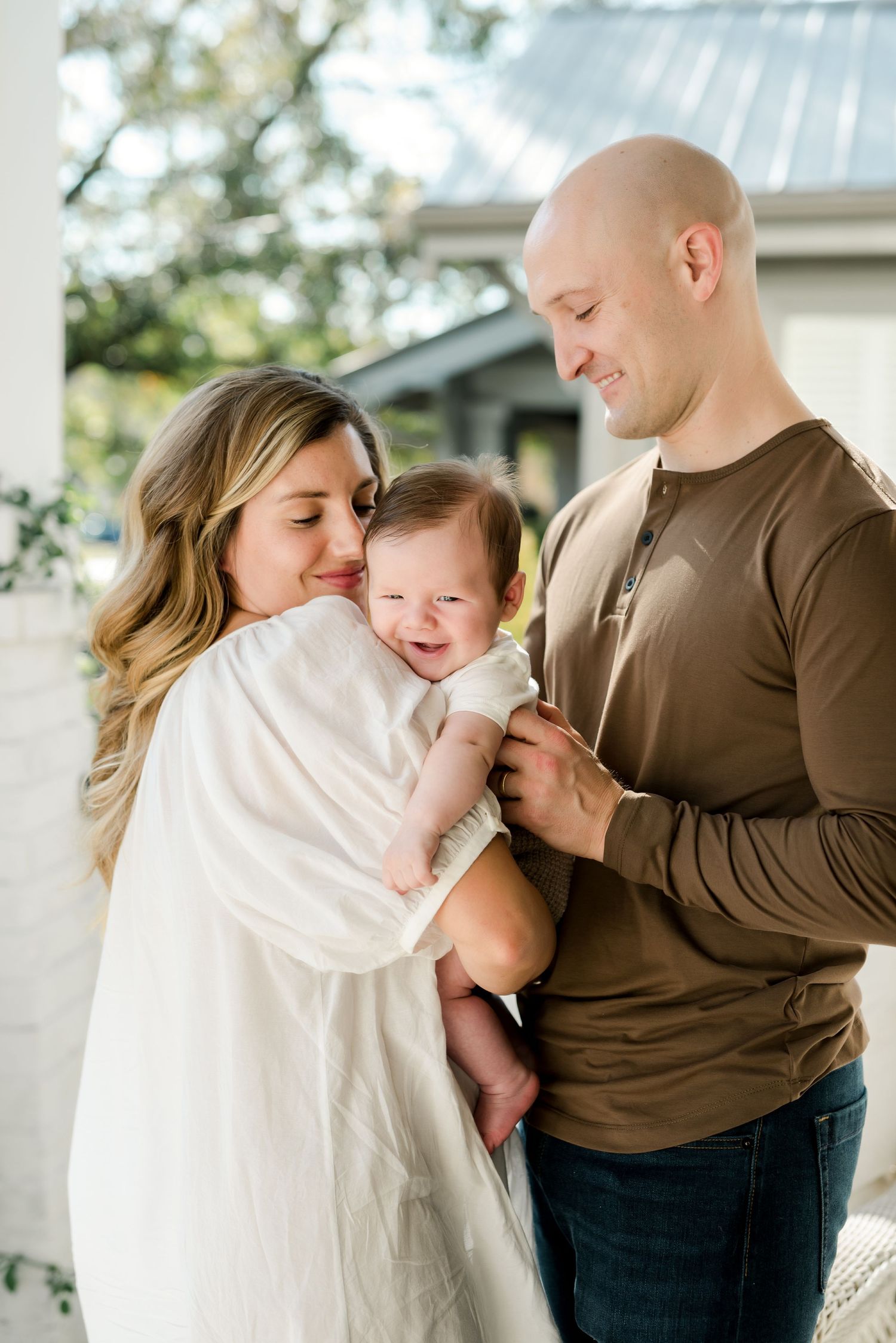 Outdoor family portrait with baby being held between parents on white porch.