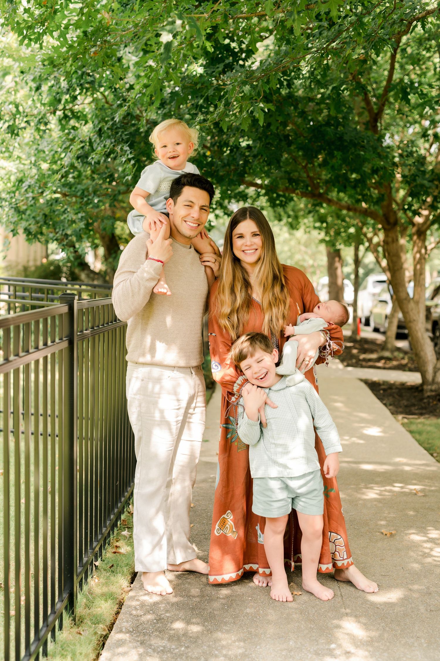 Happy family of five posing outdoors on a sunny sidewalk surrounded by lush green trees.