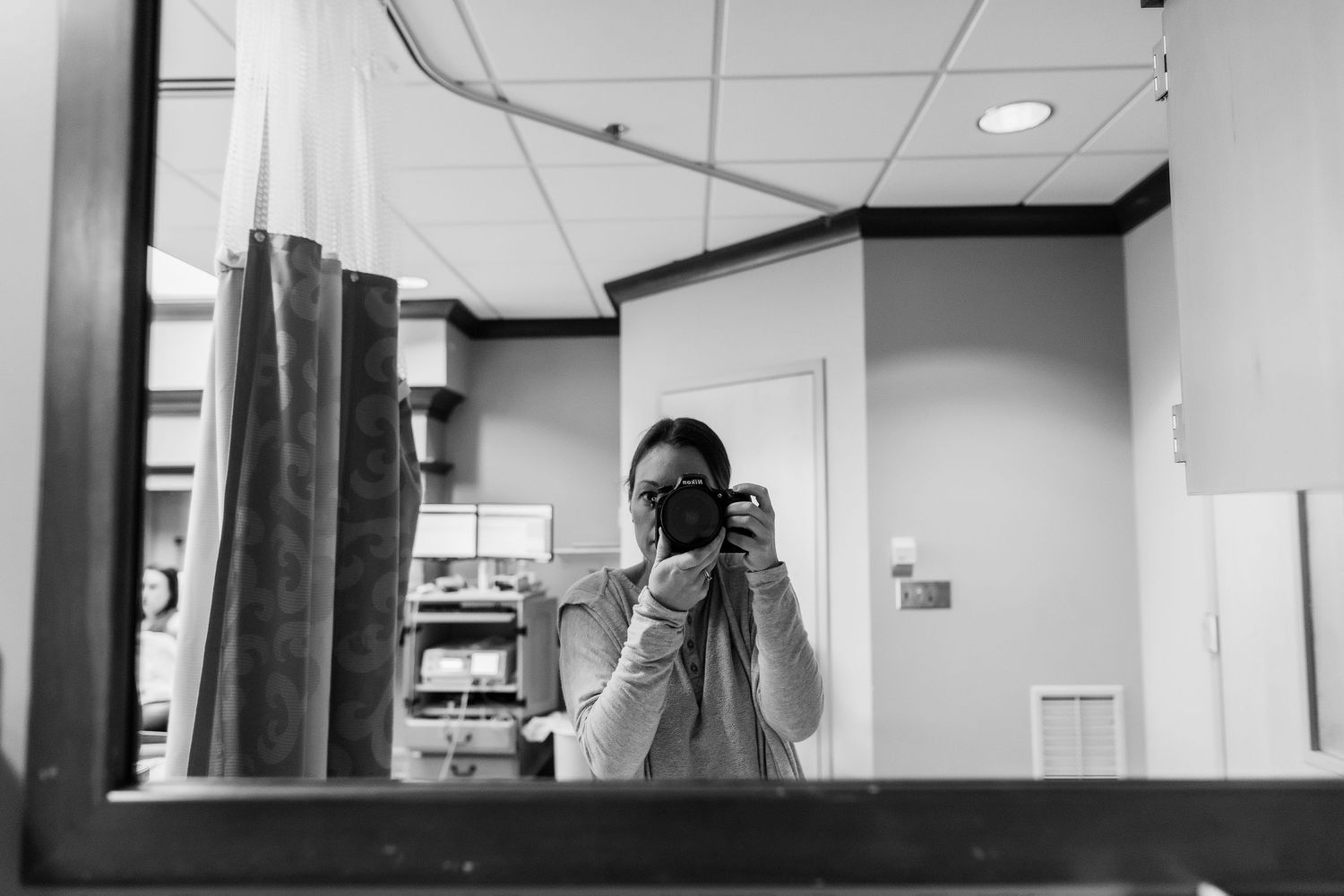 A black and white selfie taken in what appears to be a medical examination room with curtains and equipment visible.