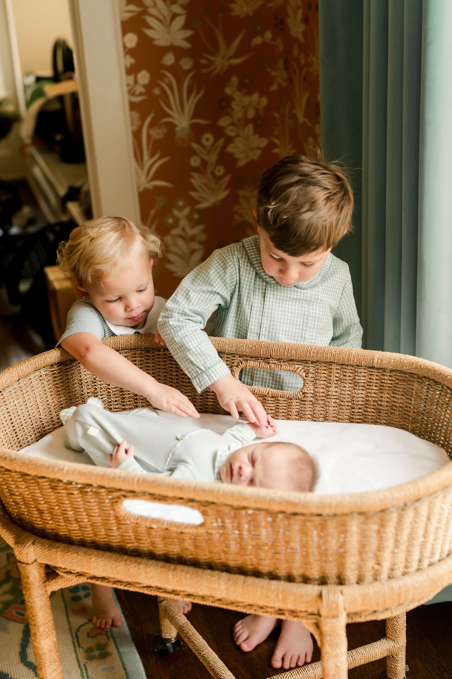 Two young siblings gently touch a swaddled newborn baby resting in a wicker bassinet.