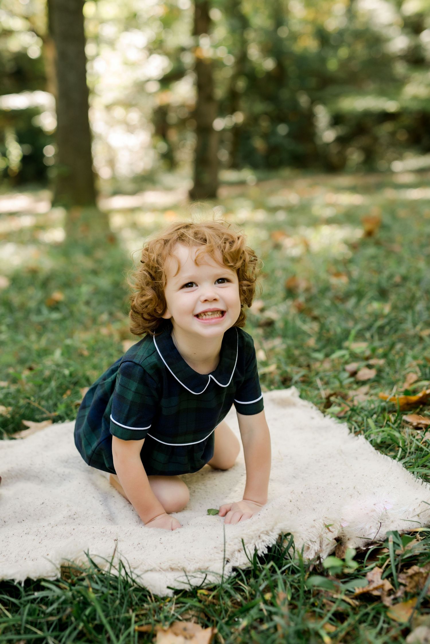 A small child in navy blue clothing crawls on a white blanket in a forest setting.