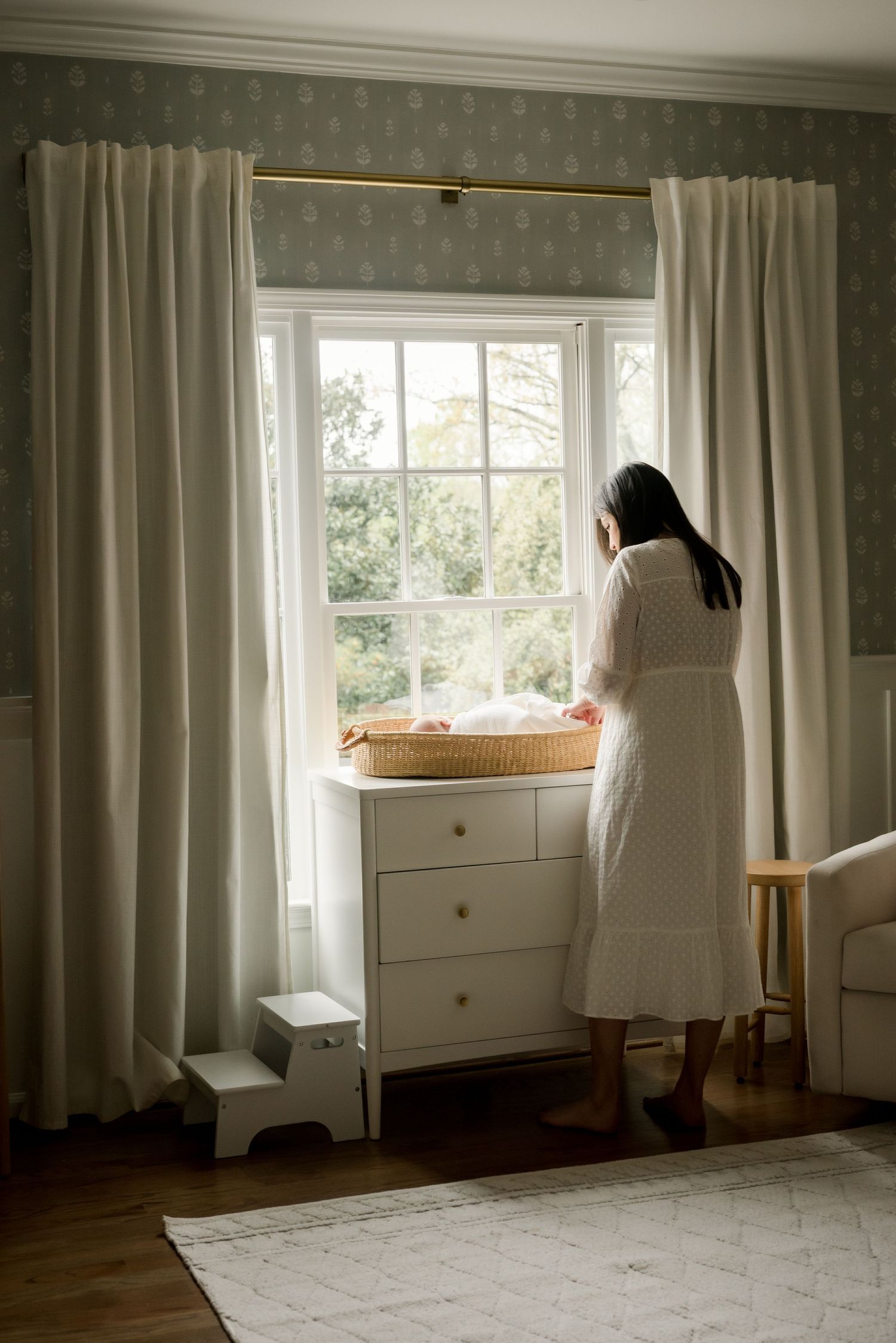 A mother in white dress looks out large window with cream curtains beside dresser and changing pad in nursery setting.