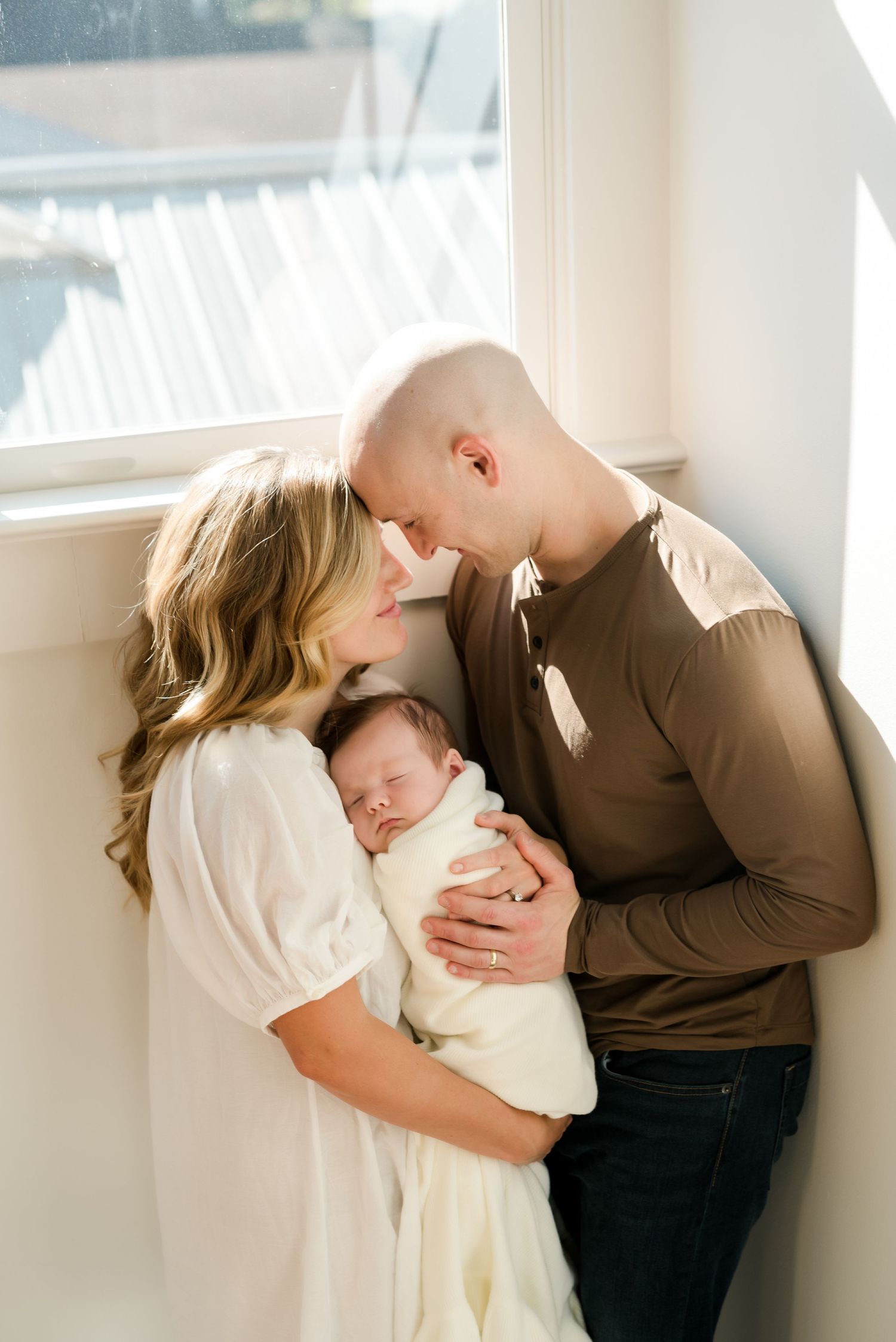 Intimate family moment as parents embrace with newborn by window.