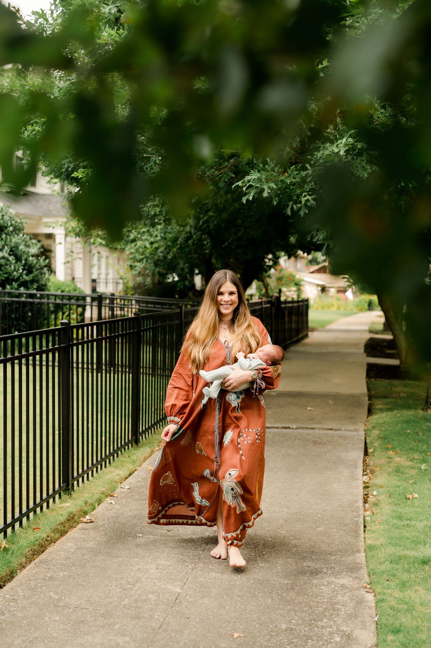 Woman in rust floral maxi dress holds newborn baby while walking on a tree-lined sidewalk.
