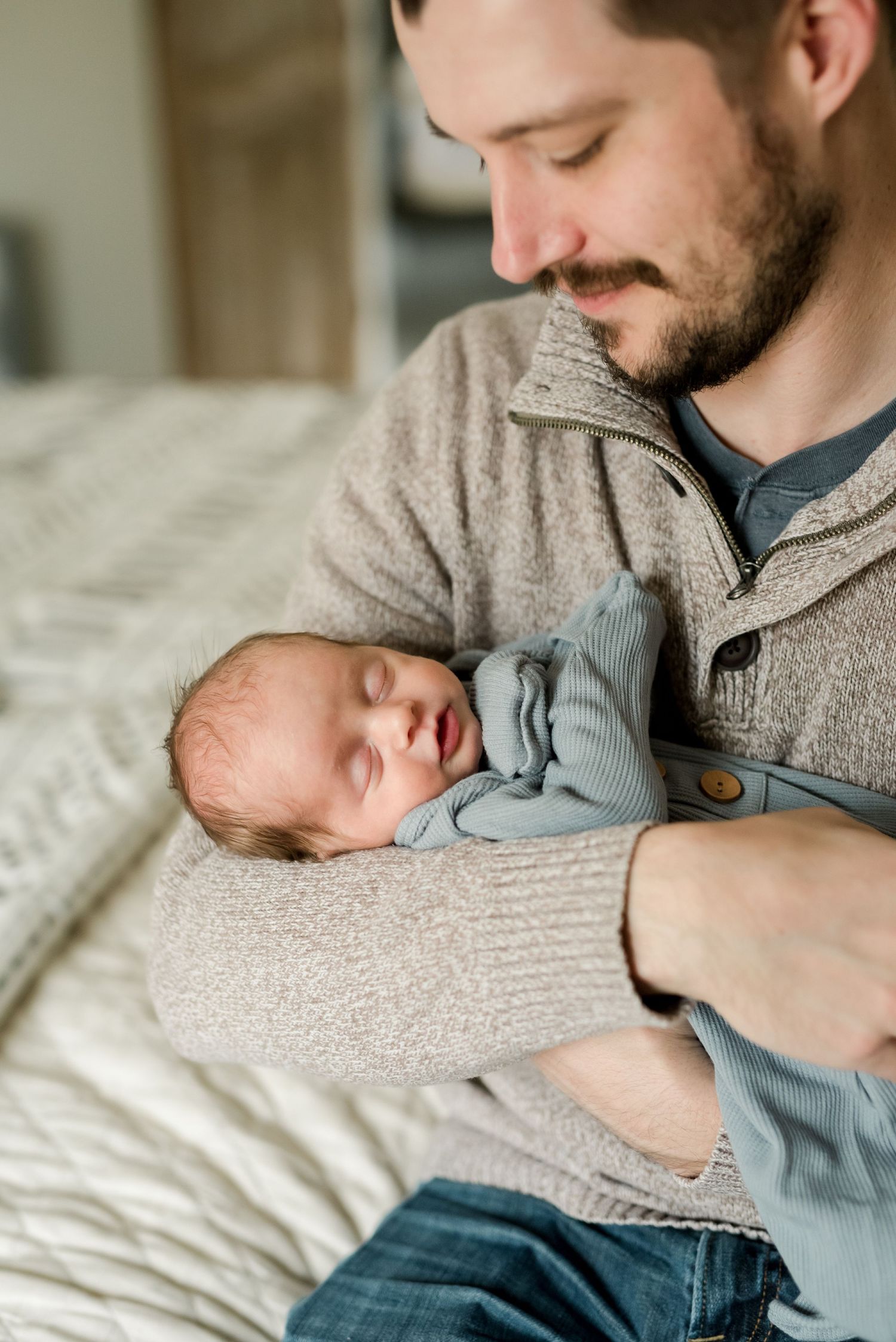 Father in grey sweater holding sleeping newborn baby dressed in blue outfit on white bedding.