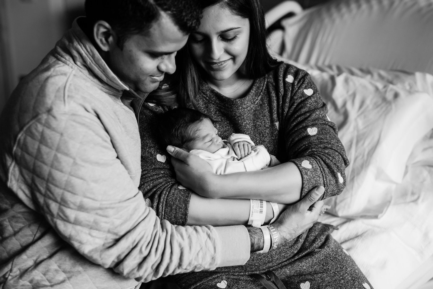 Black and white photo of a tender moment with parents cradling their newborn baby on a cozy bed.