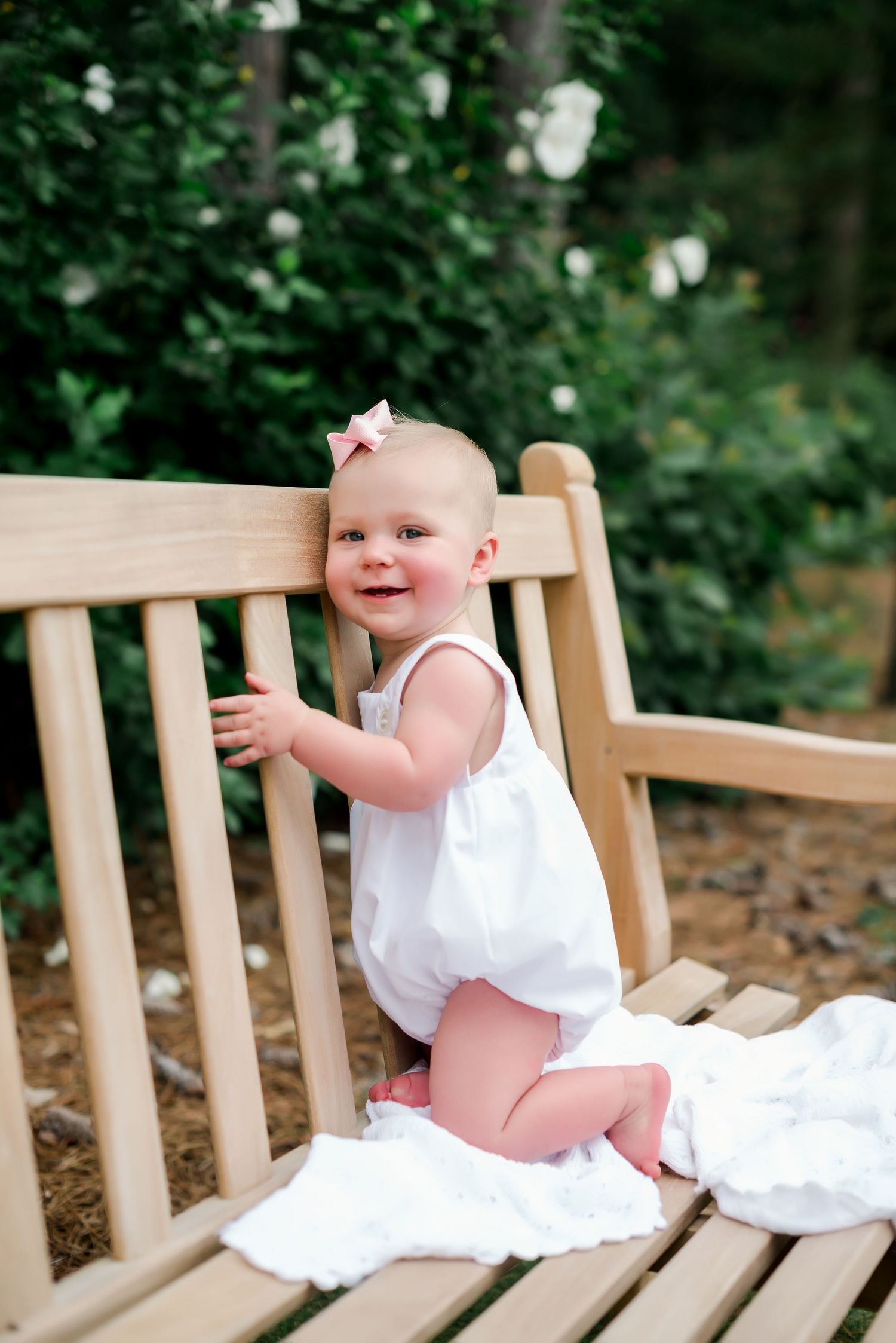 Smiling baby in white outfit plays on wooden garden bench surrounded by greenery.