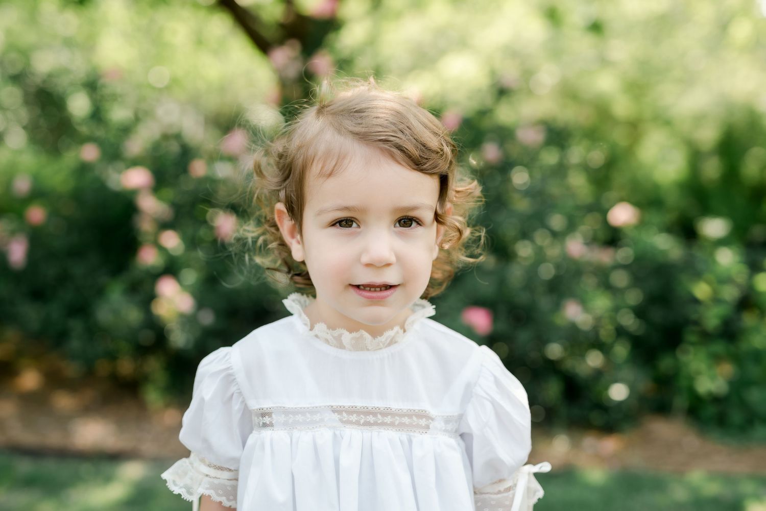 Young person in white dress stands outdoors among blooming garden flowers.