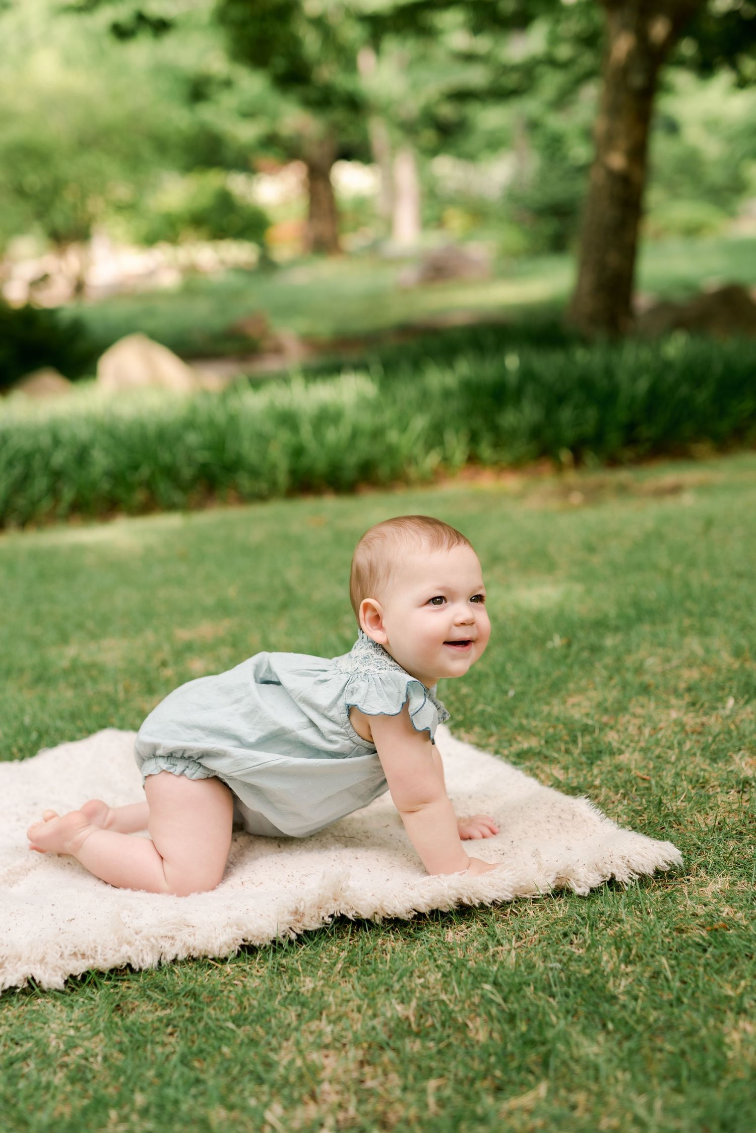 Baby crawling on soft blanket in garden with trees and grass in background.
