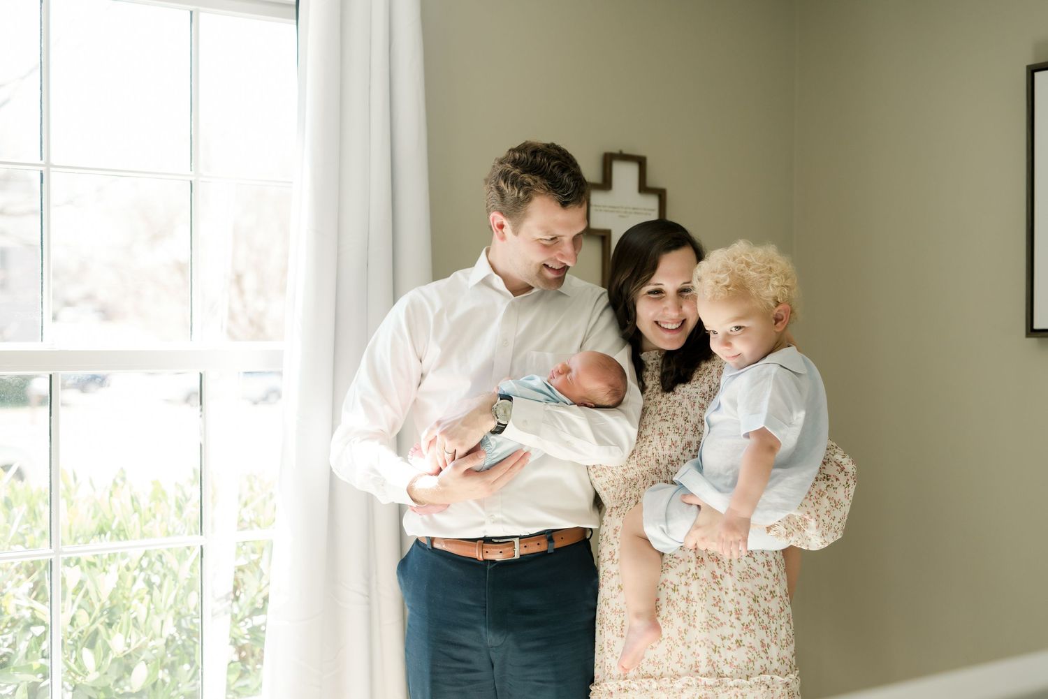 Family standing by window with toddler and newborn baby in casual home setting.