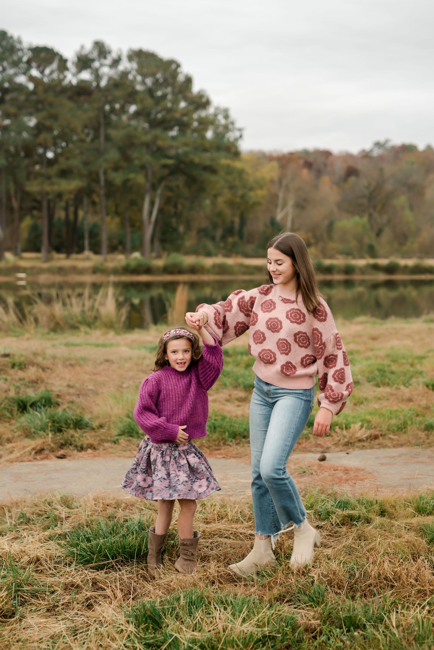 Two people in cozy fall sweaters walk together in a scenic field by a lake during autumn.