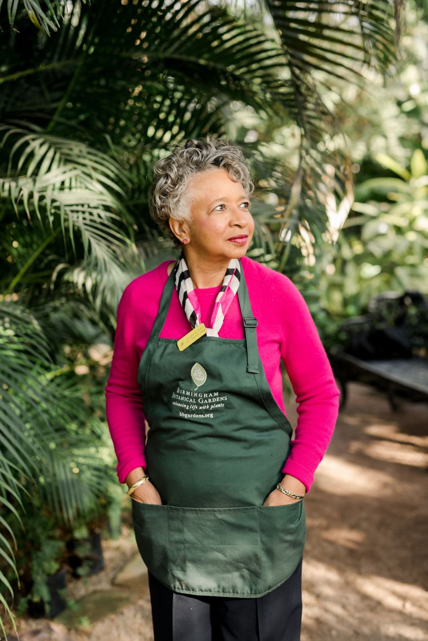 Person in a green apron and pink sweater poses cheerfully in front of tropical plants.