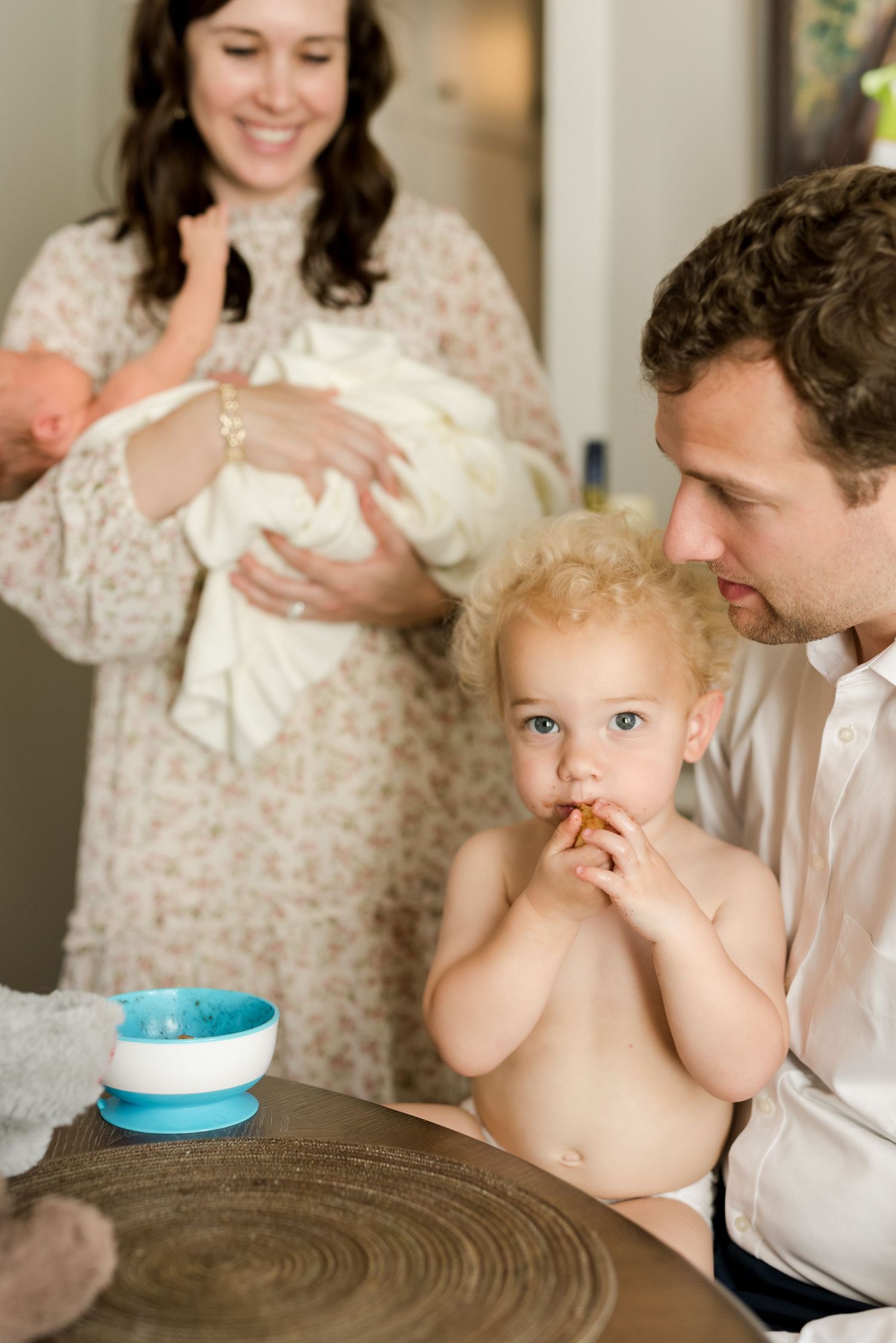 Family mealtime scene with shirtless toddler eating while parents hold newborn.