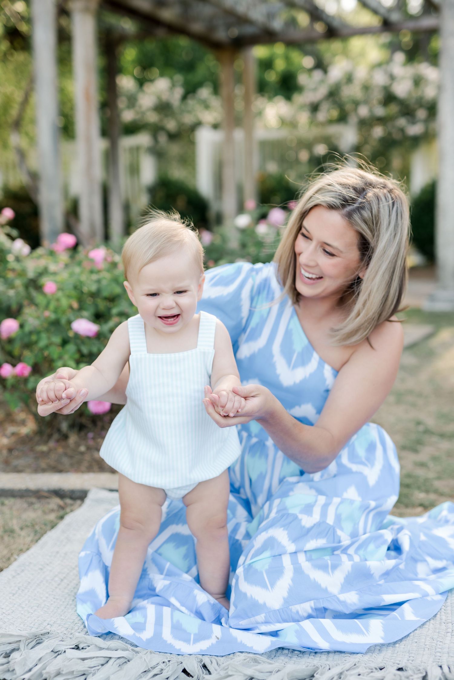 Mother and baby in matching blue and white outfits sitting on blanket in garden.
