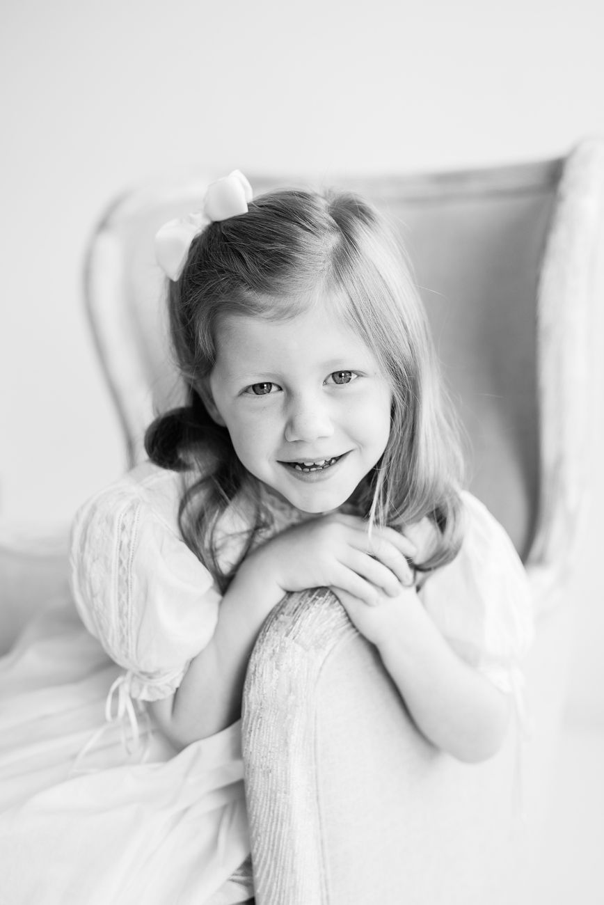 A black and white portrait of a child in white dress sitting in an elegant chair.