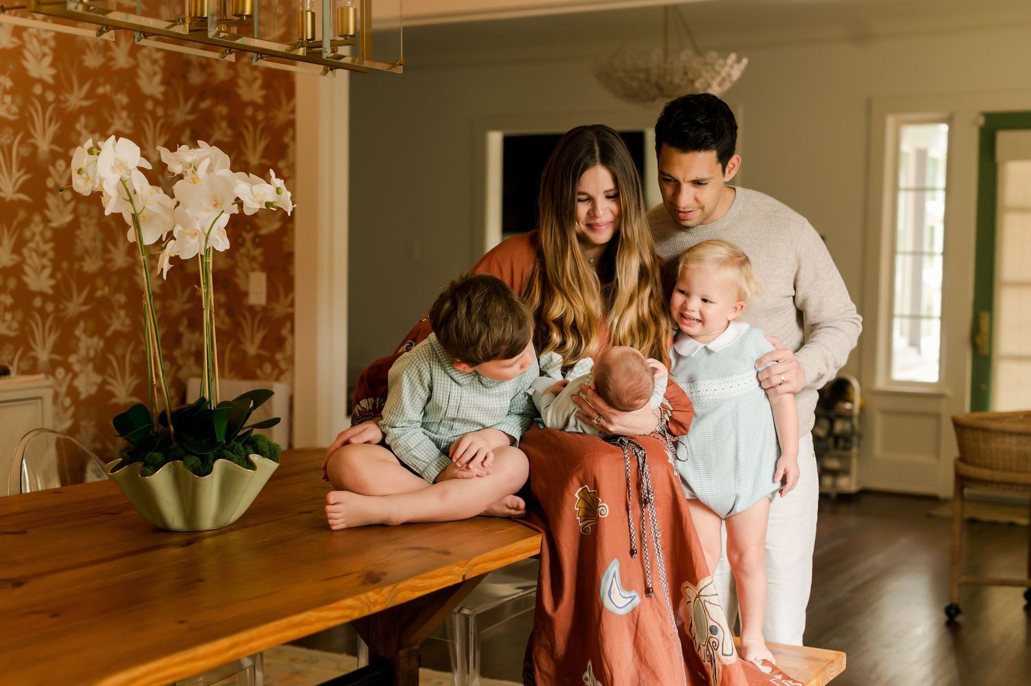 A young family of five gathers around a wooden dining table in a warmly decorated home interior.