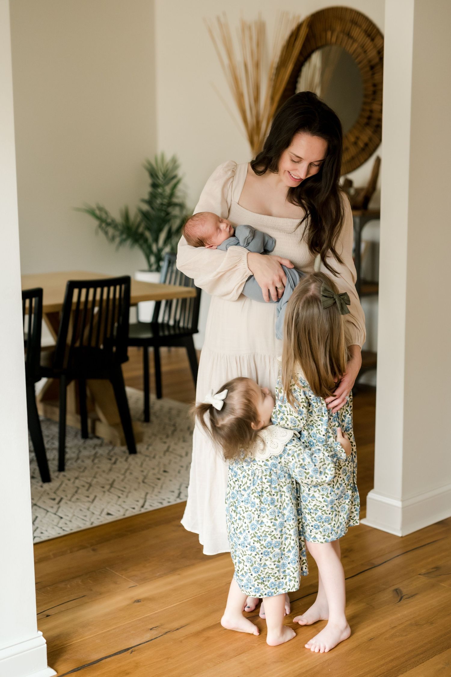 Family moment at home with young child in floral dress and baby while standing near dining area.