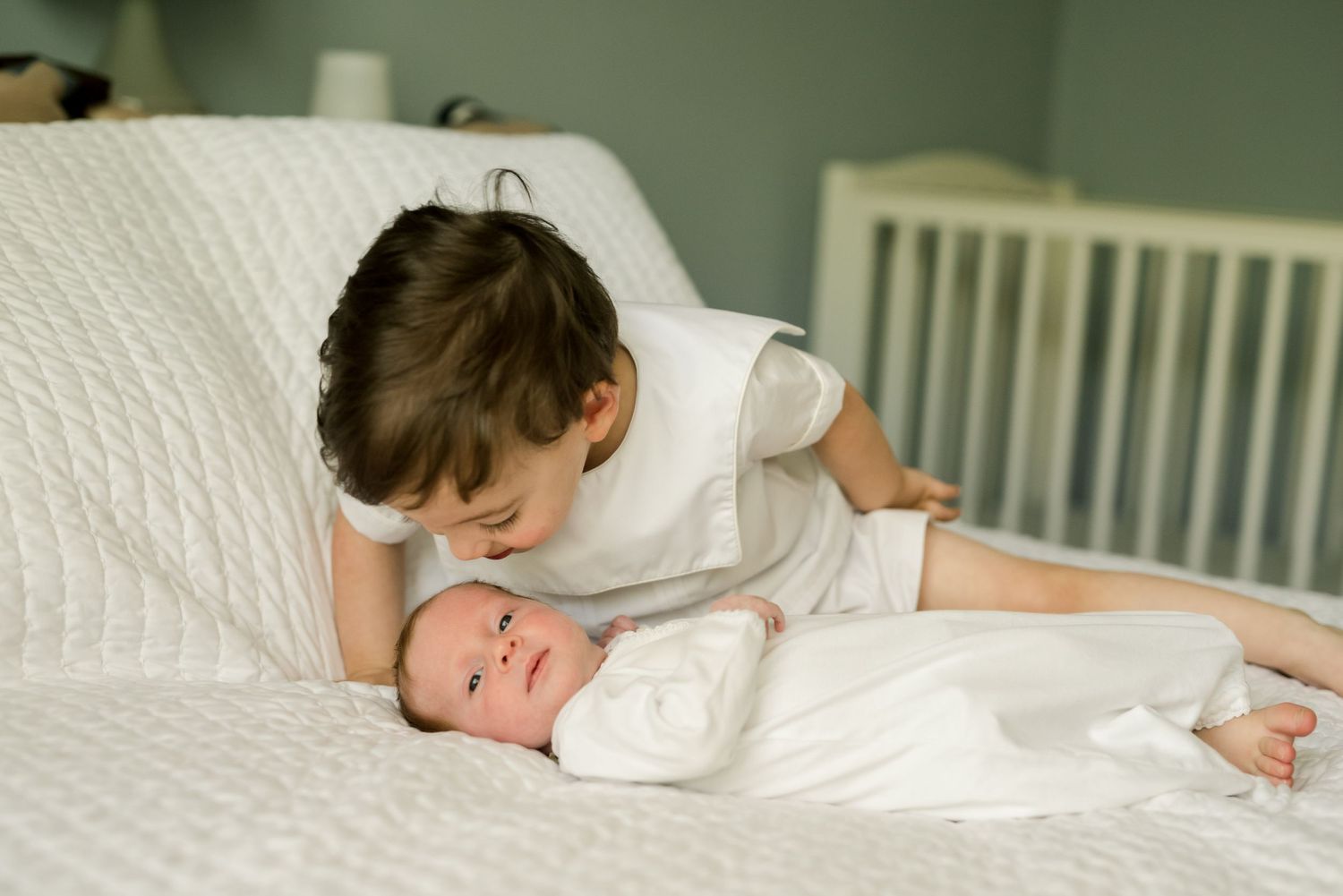 Sibling bonding moment as older child leans over to look at baby on white bedding.