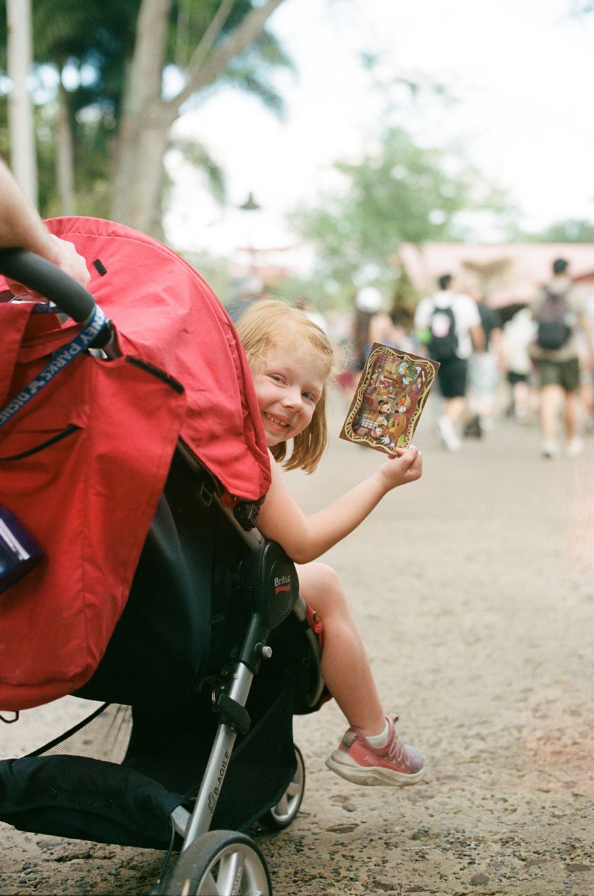 A child peeks out from behind a red stroller while holding a phone in a crowded park.