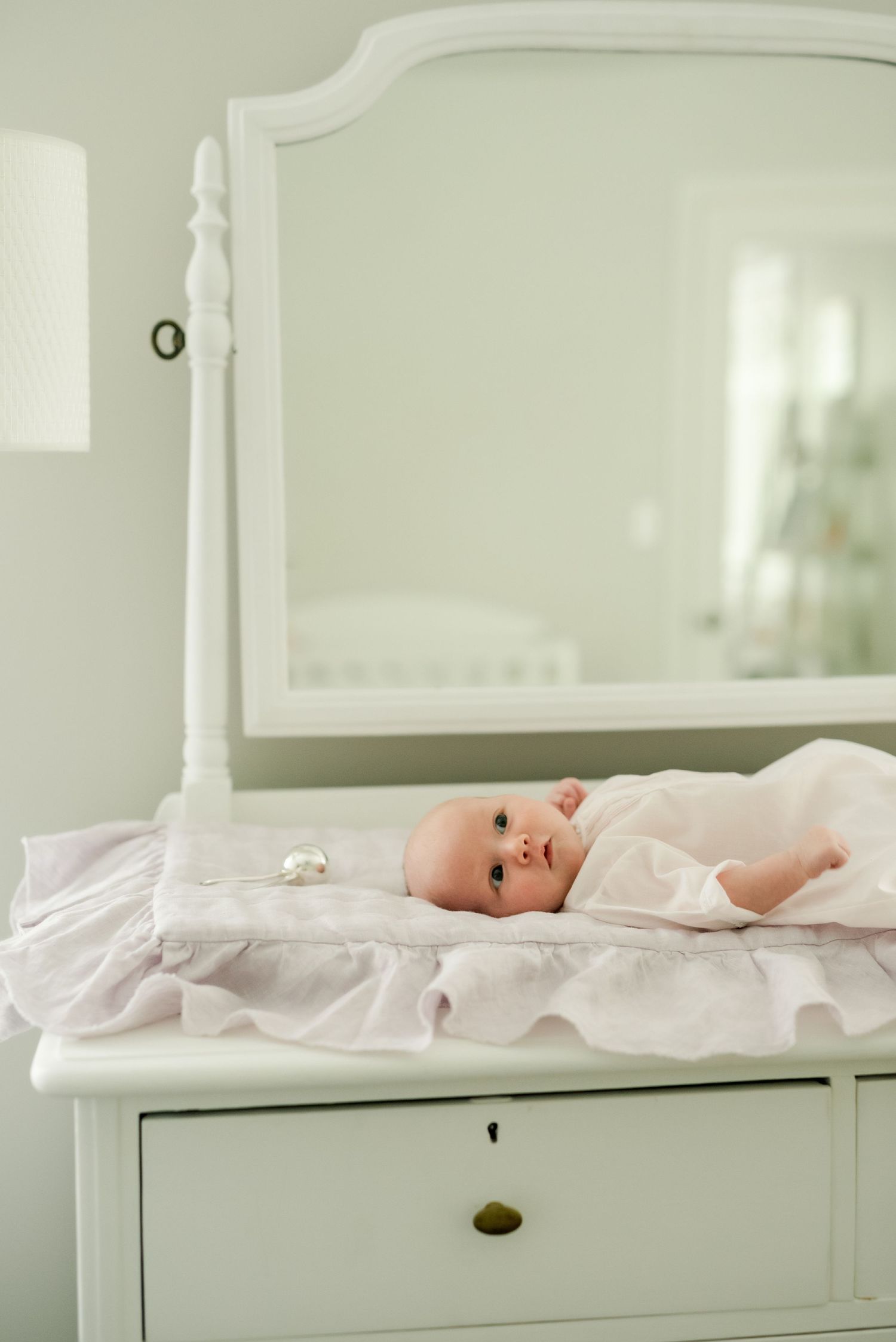Baby in white outfit lying on vintage white changing table with mirror.