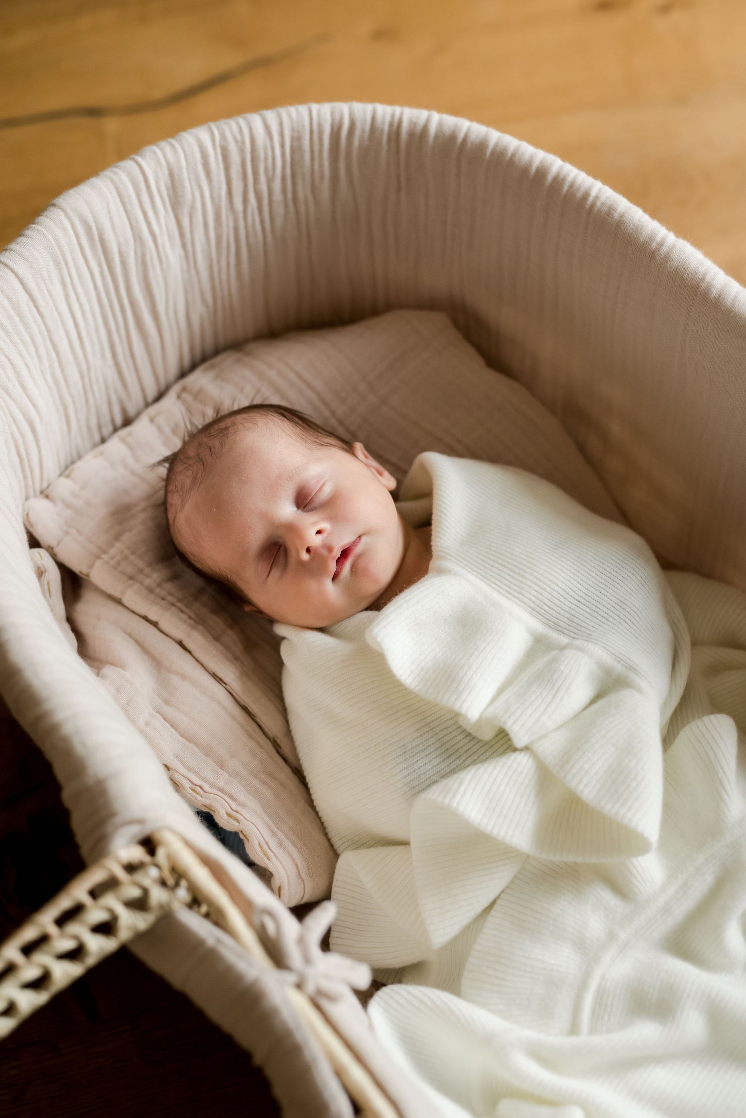 Sleeping newborn baby in white outfit lying in cream-colored bassinet.